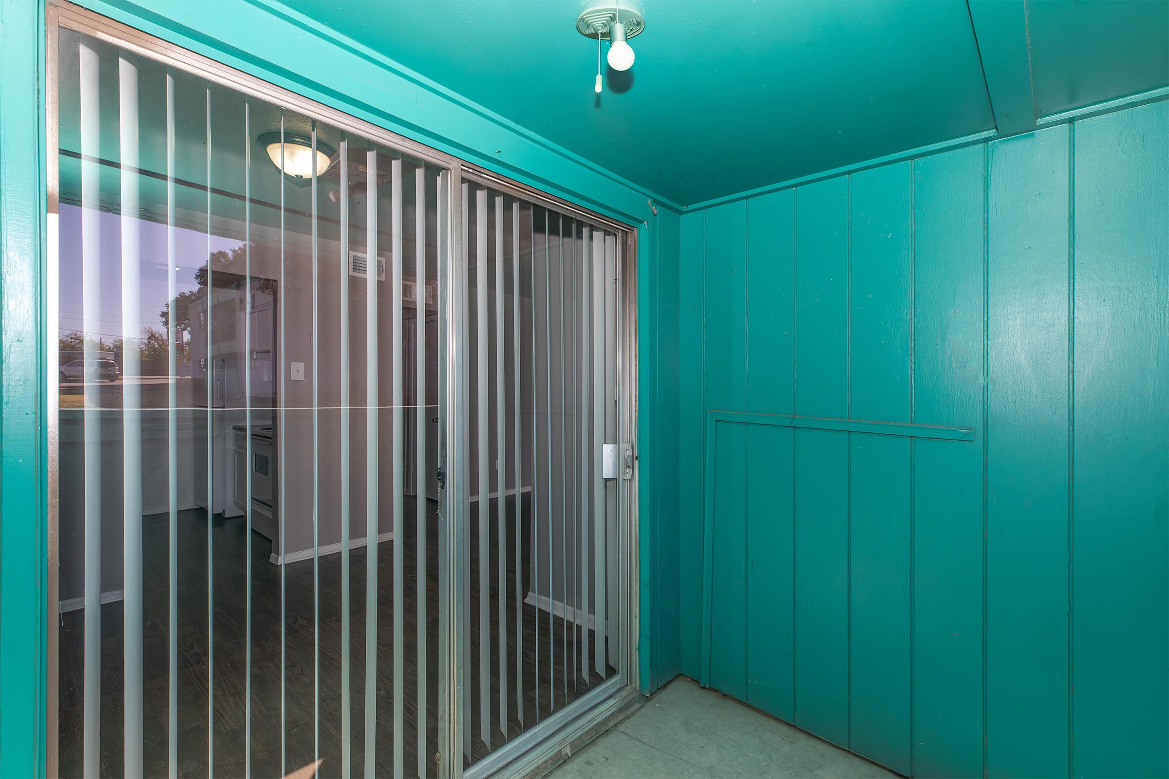 Interior view of a room with teal-colored walls. A sliding glass door with vertical blinds provides access to the outside. The floor is dark, possibly wood or laminate. Natural light filters in from the door, and there is a light fixture on the ceiling.