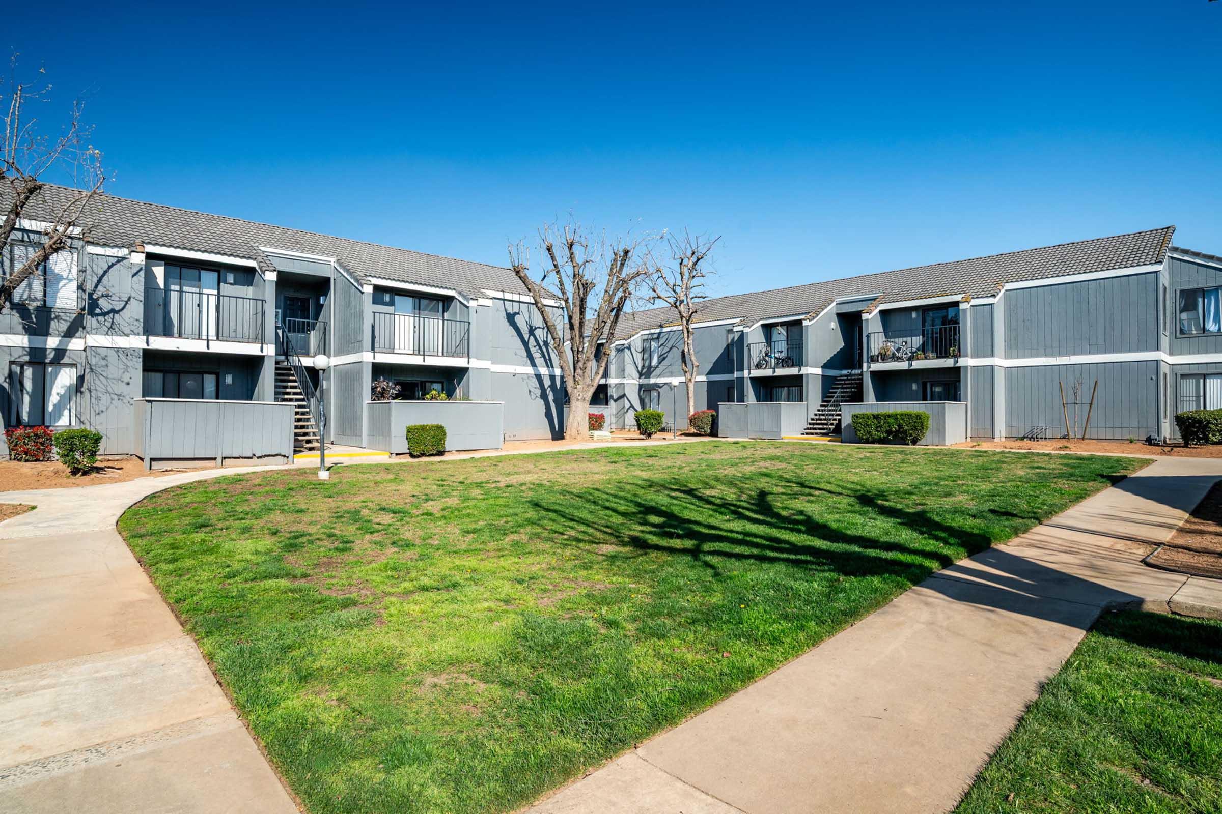 A view of a multi-unit apartment complex with gray exterior, featuring two-story buildings arranged around a grassy courtyard. There are pathways leading through the lawn, and a few trees are visible, providing shade in the area. The sky is clear and blue, indicating a sunny day.