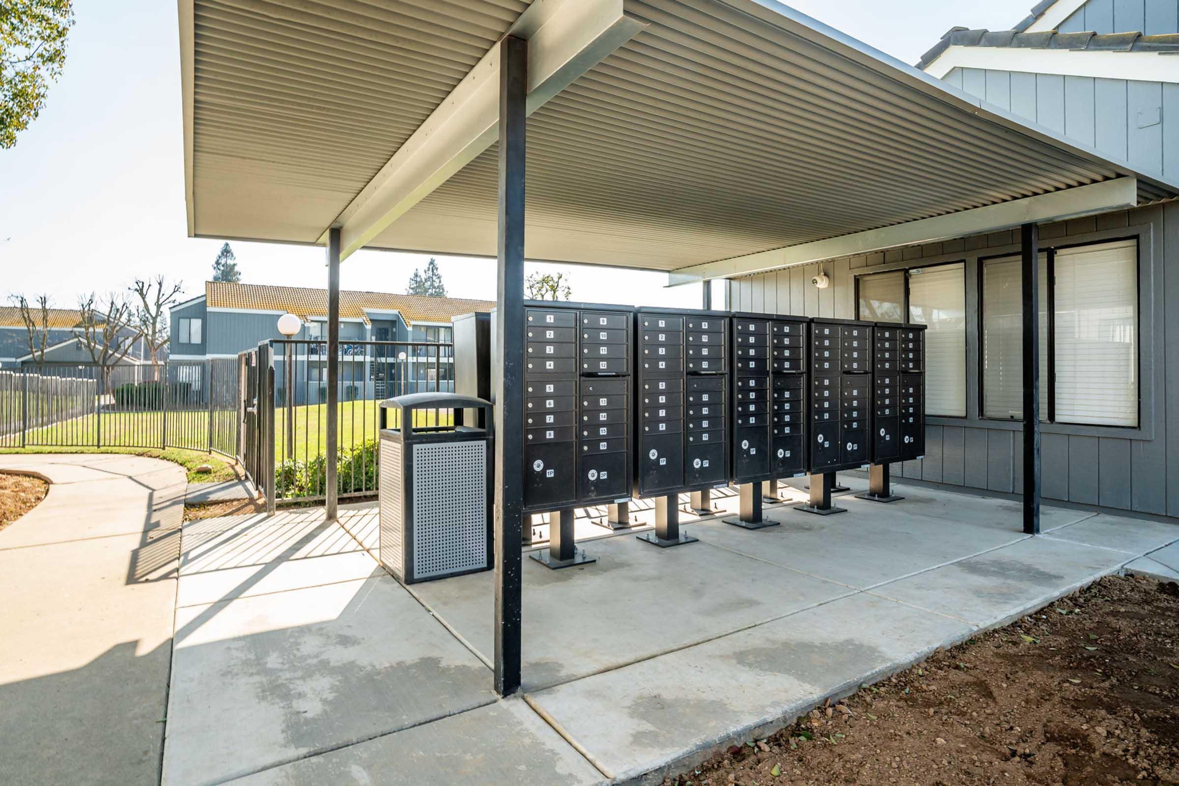 A row of locked mailboxes under a covered area beside a building. There is a trash bin nearby and green grass visible in the background, along with other buildings. The setting is well-lit and tidy, suggesting a residential or community housing area.