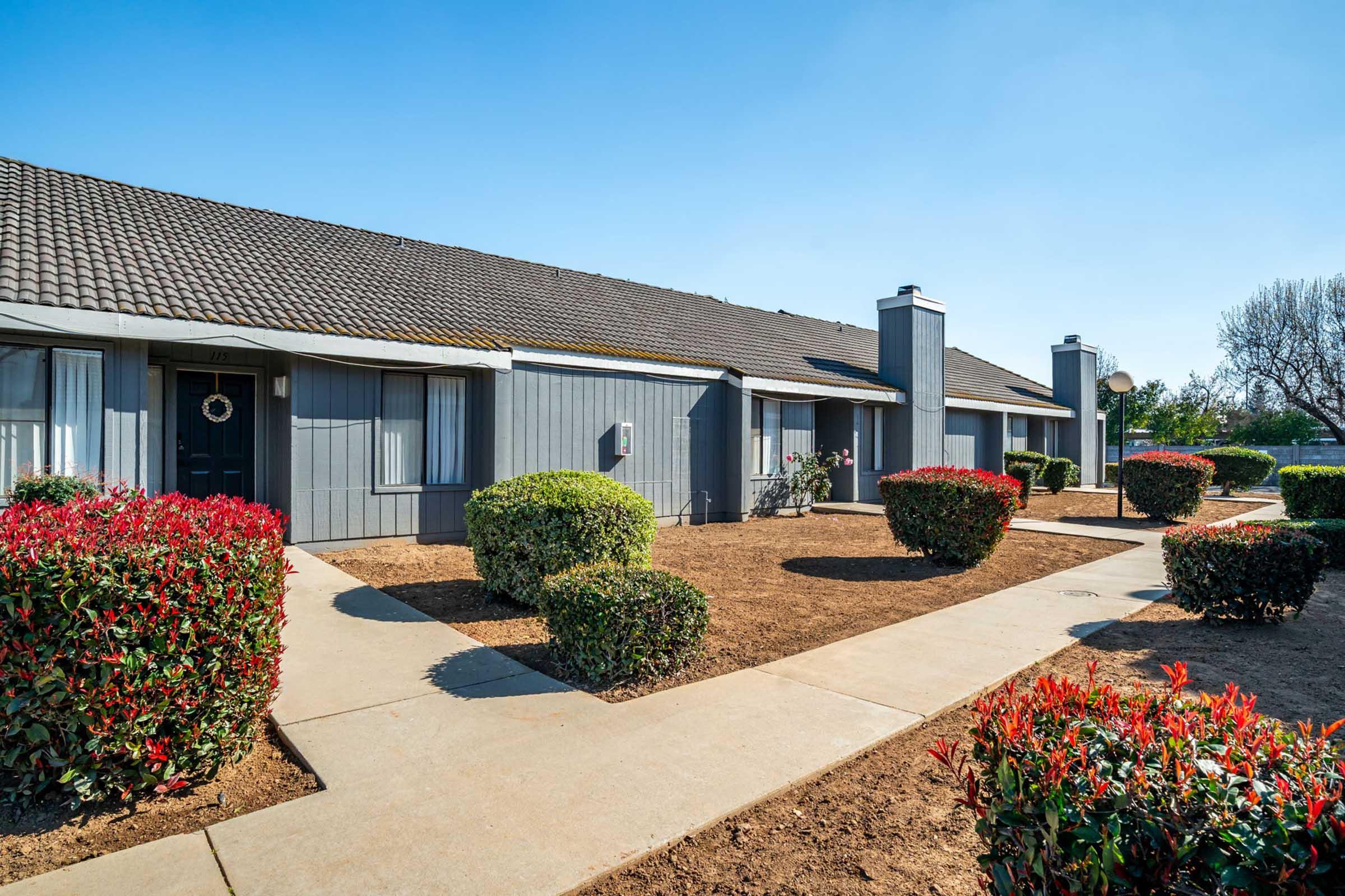 A well-maintained residential building with a gray exterior and a sloped roof. The front features neatly trimmed bushes in vibrant colors, a pathway leading to the entrance, and a clear blue sky above. The landscape is tidy, contributing to a welcoming atmosphere.
