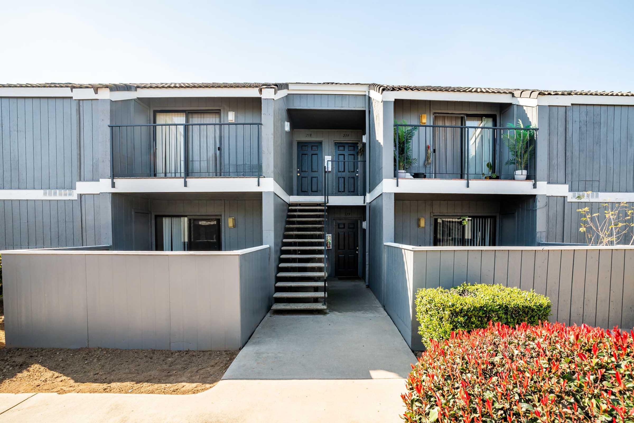 A two-story apartment building featuring a gray exterior with a central staircase leading to the upper floor. Each unit has a balcony with potted plants, and there are manicured bushes and landscaped areas at the front. The sky is clear and blue, indicating a sunny day.