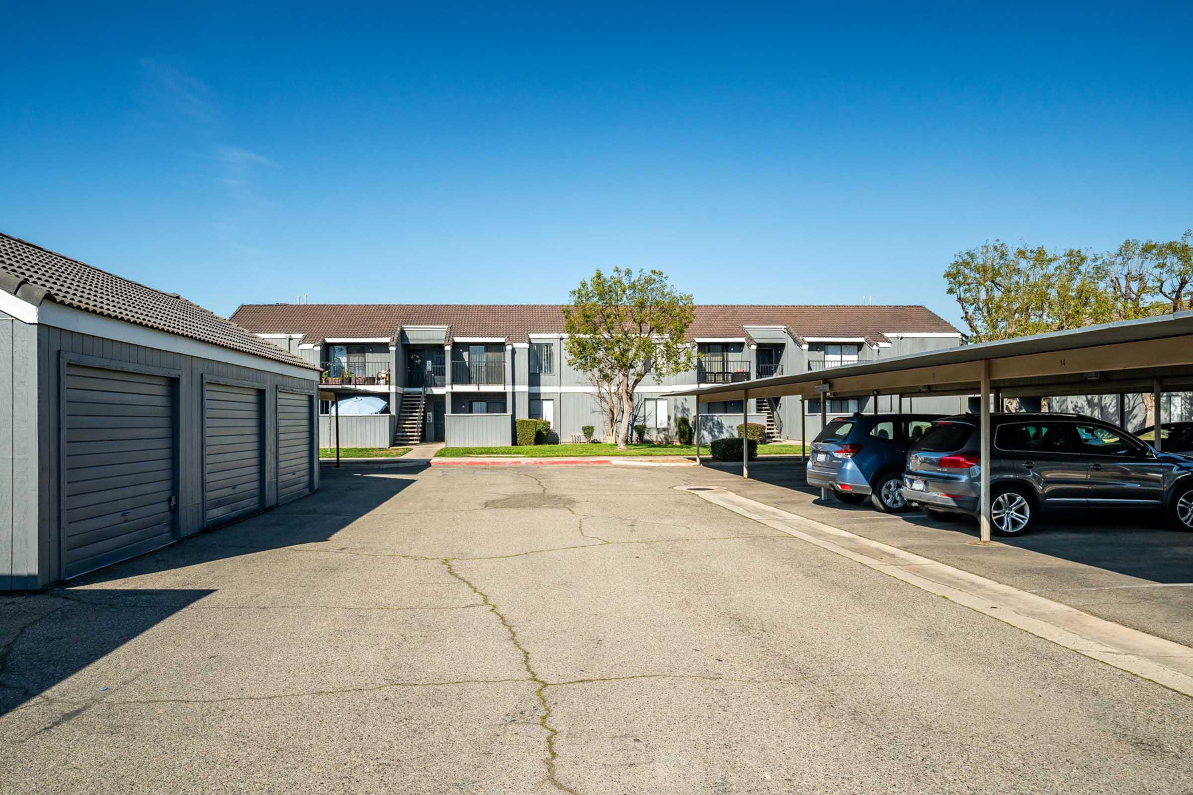 A view of an apartment complex featuring two-story buildings, a parking area with covered and uncovered spaces, and a clear blue sky. Trees are visible in the background, and there are several parked cars along the driveway and under the awning.