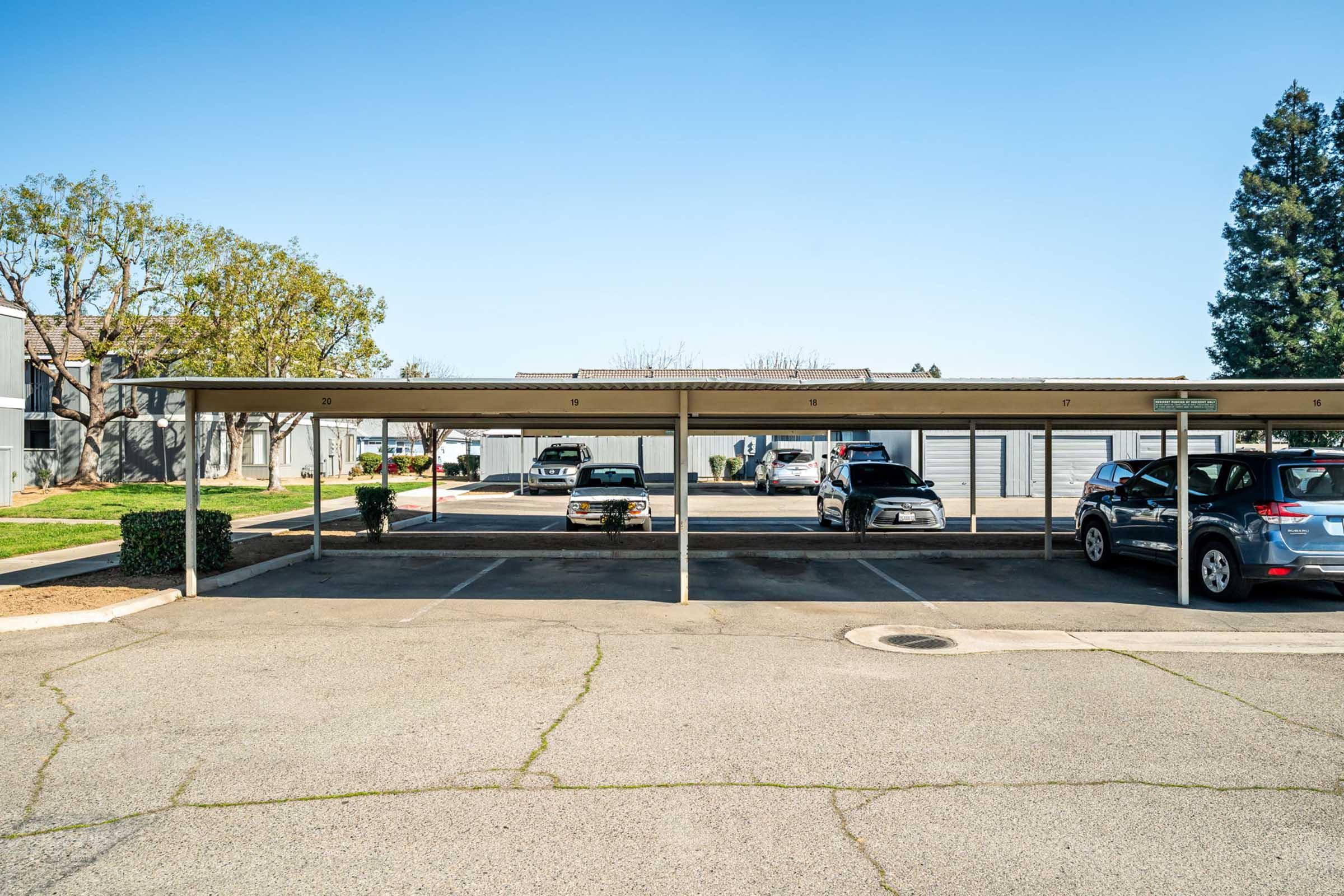 A parking lot with several covered spaces, featuring a few parked cars. The background includes a tree and a clear blue sky. The area is well-maintained, and the pavement shows clear parking lines.