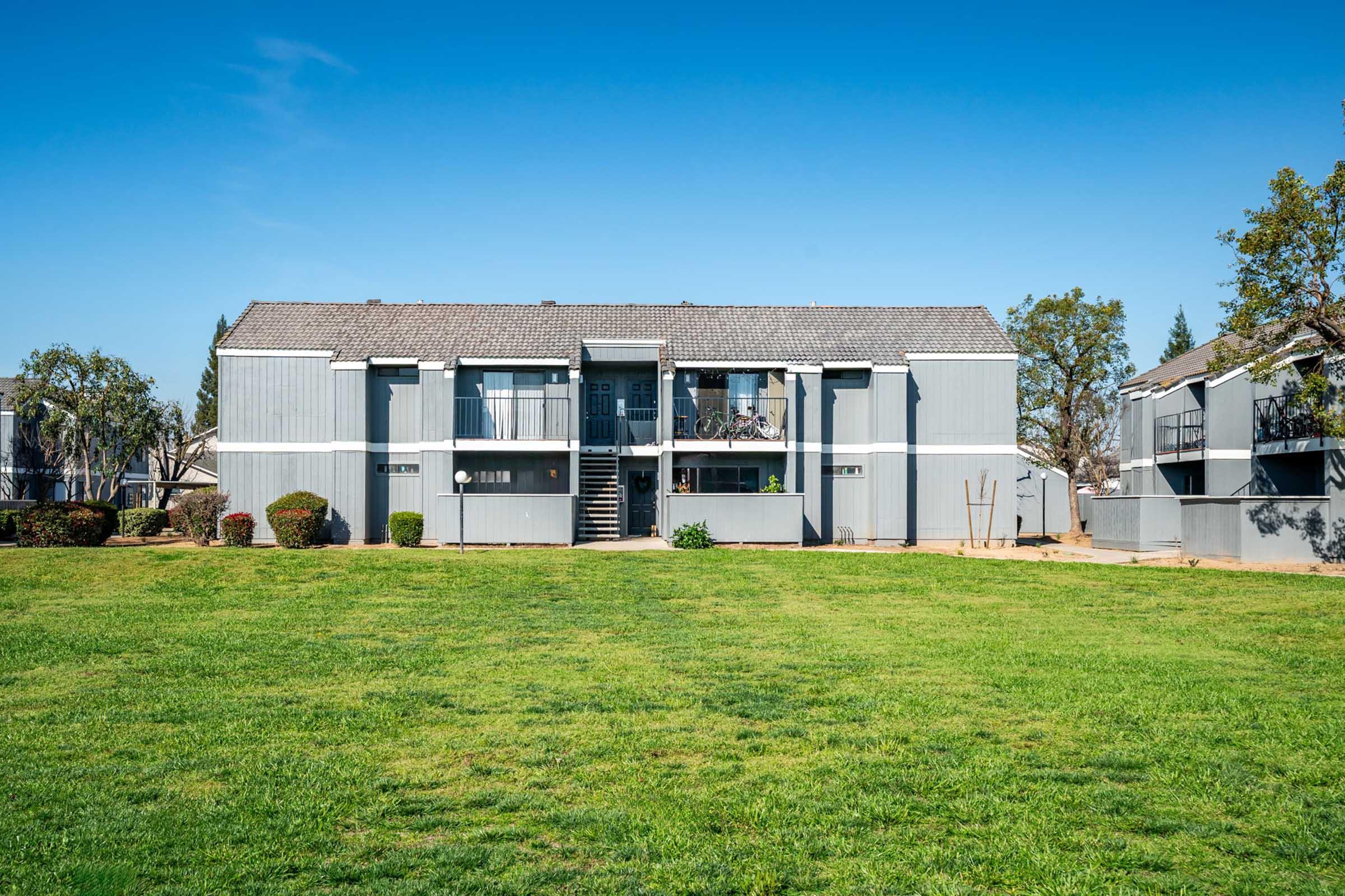 Two-story apartment building with gray siding, featuring balconies on the upper floor. The grass in front is well-maintained, with a few shrubs on either side. Clear blue sky above, suggesting a sunny day. A spacious area around the building is visible, possibly part of a residential complex.