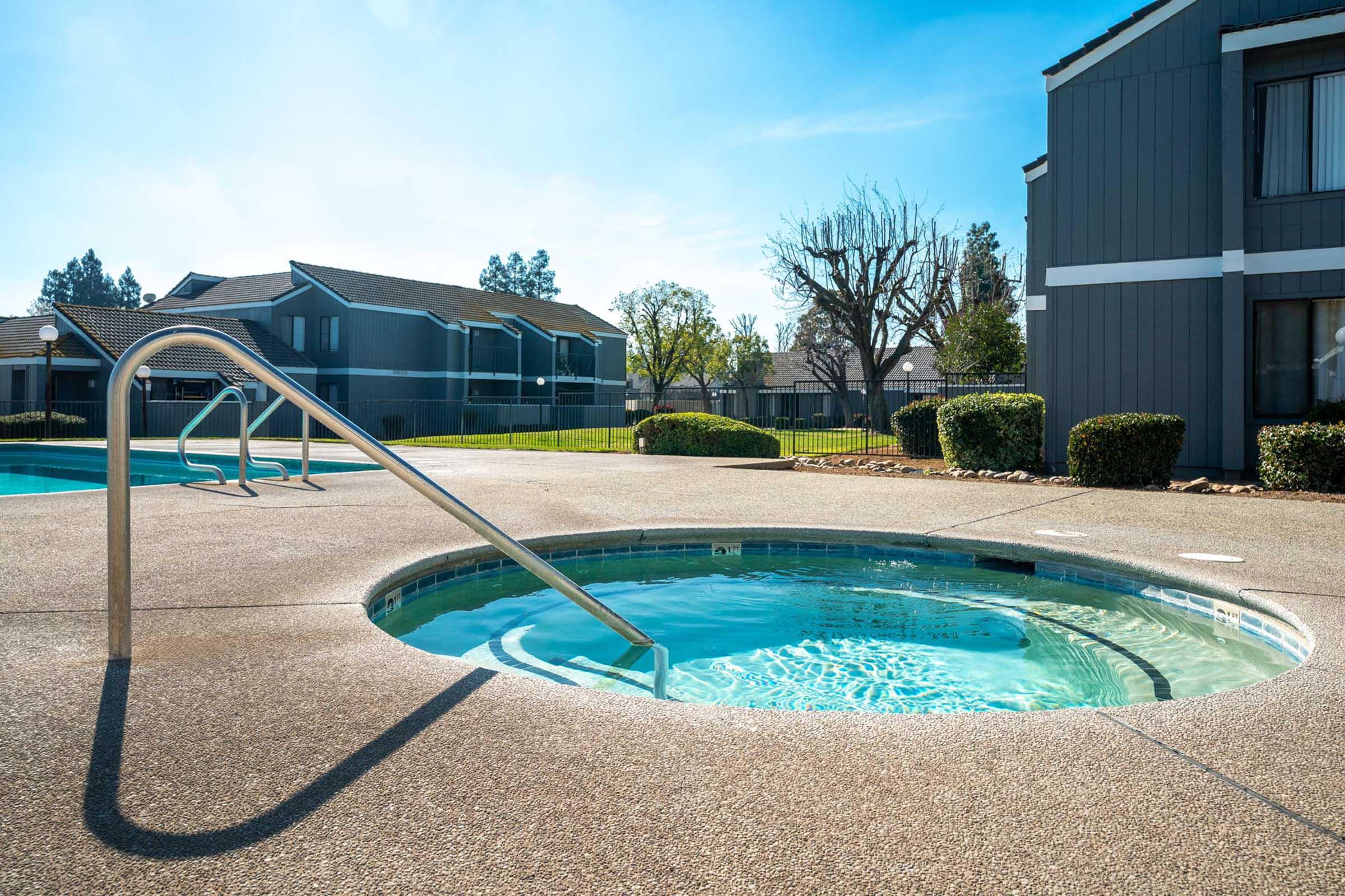 A clear hot tub surrounded by a textured concrete deck, with a handrail for support. Residential buildings in the background are partially visible, and the scene is bathed in natural sunlight, with a few trees and a pool area also in view. The atmosphere is calm and inviting.