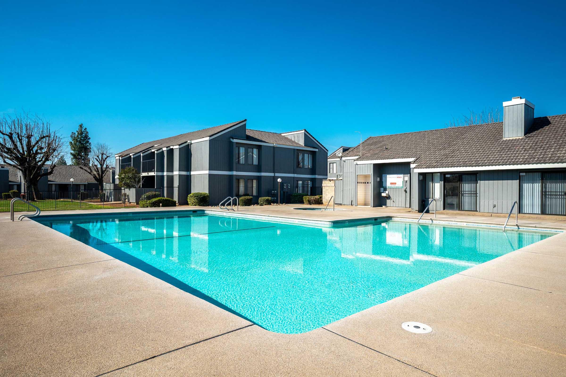 A clear blue swimming pool surrounded by concrete pathways, with lounge chairs and grassy areas nearby. In the background, there are two grey apartment buildings under a bright blue sky, creating a serene and inviting atmosphere.
