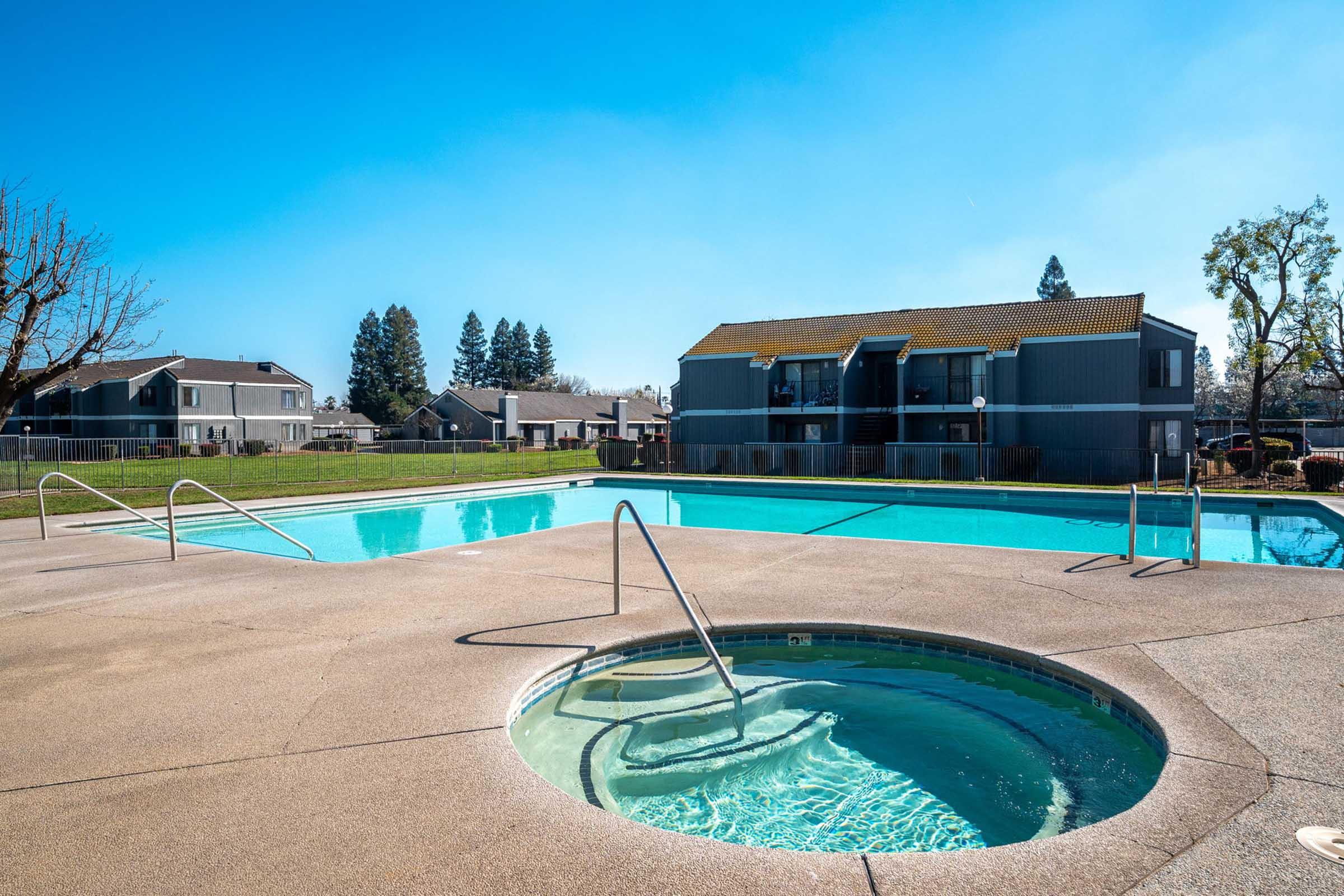 A bright, sunny day at an apartment complex featuring a swimming pool and hot tub. The pool is clear and inviting, surrounded by a well-maintained lawn and trees. In the background, there are residential buildings, creating a relaxing community atmosphere.