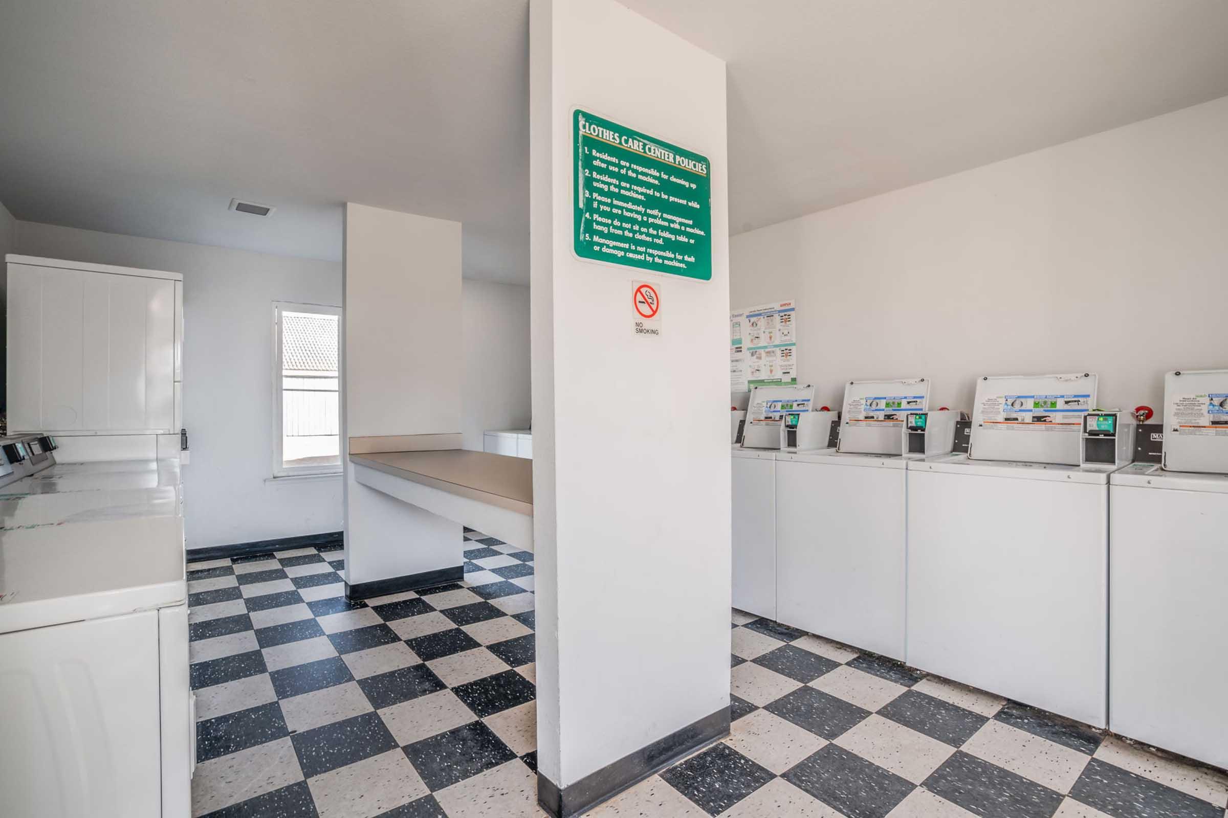 A clean and bright laundry room featuring several white washing machines and dryers arranged along one wall. There is a folding station with a countertop in the center. The floor is black and white checkered, and a green sign providing laundry instructions is mounted on the wall.