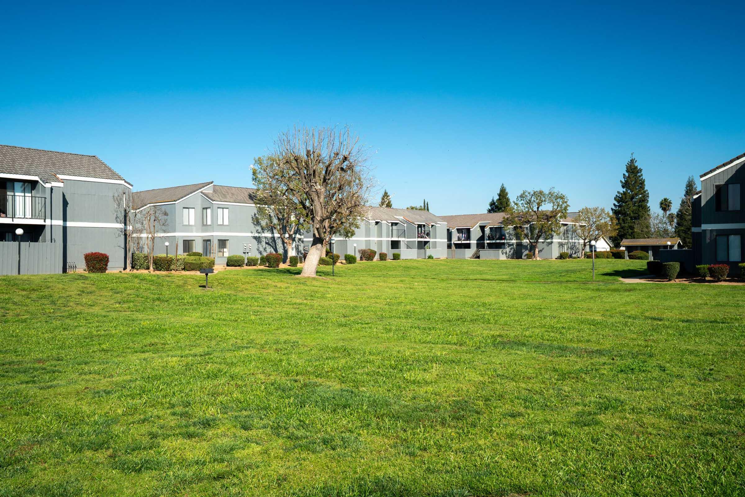 A view of a residential apartment complex featuring two-story buildings with gray exteriors, surrounded by well-maintained green lawns and a few trees under a clear blue sky.