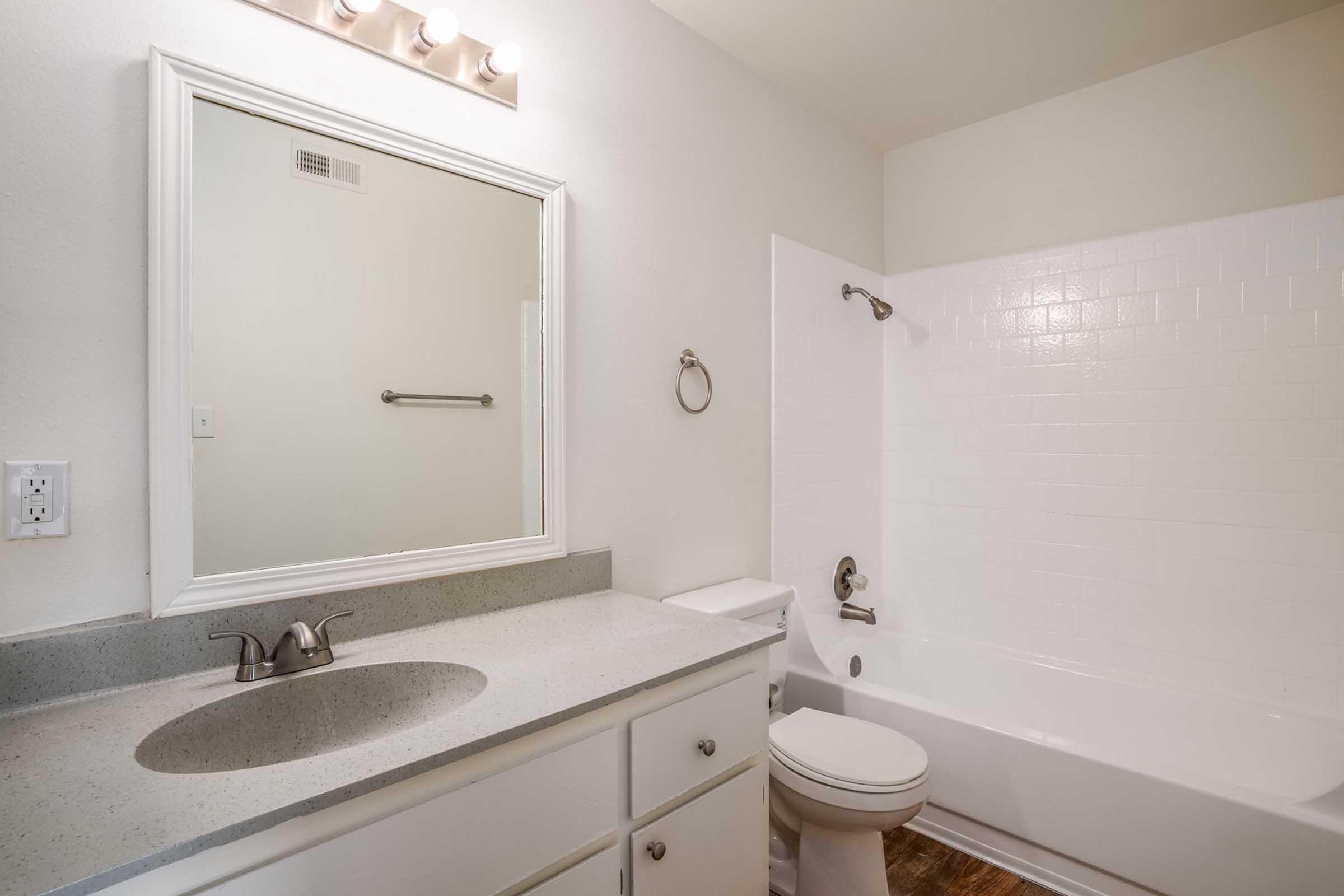 A clean, modern bathroom featuring a white tub with a shower, a rectangular mirror above a light-colored countertop, and a simple vanity with storage below. The walls are painted light, and there is a towel rack on the wall next to the mirror.