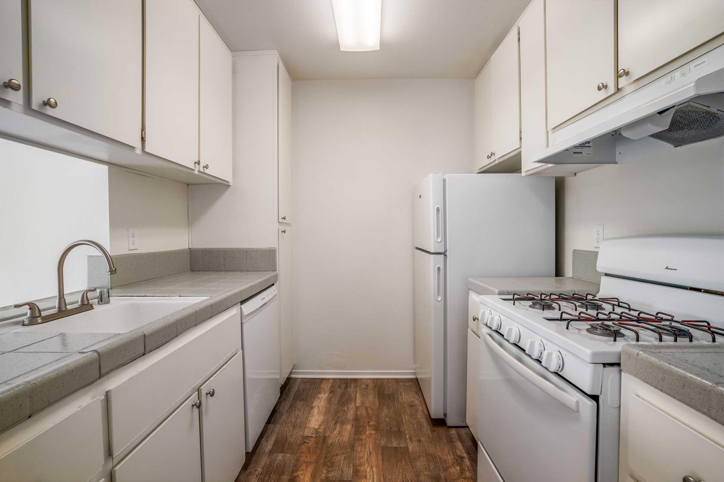 A compact kitchen featuring white cabinets, a dual-basin sink, a refrigerator, and a gas stove. The countertops are light gray, and the flooring is a brown wood-like material. The kitchen has bright overhead lighting and a clean, modern design.
