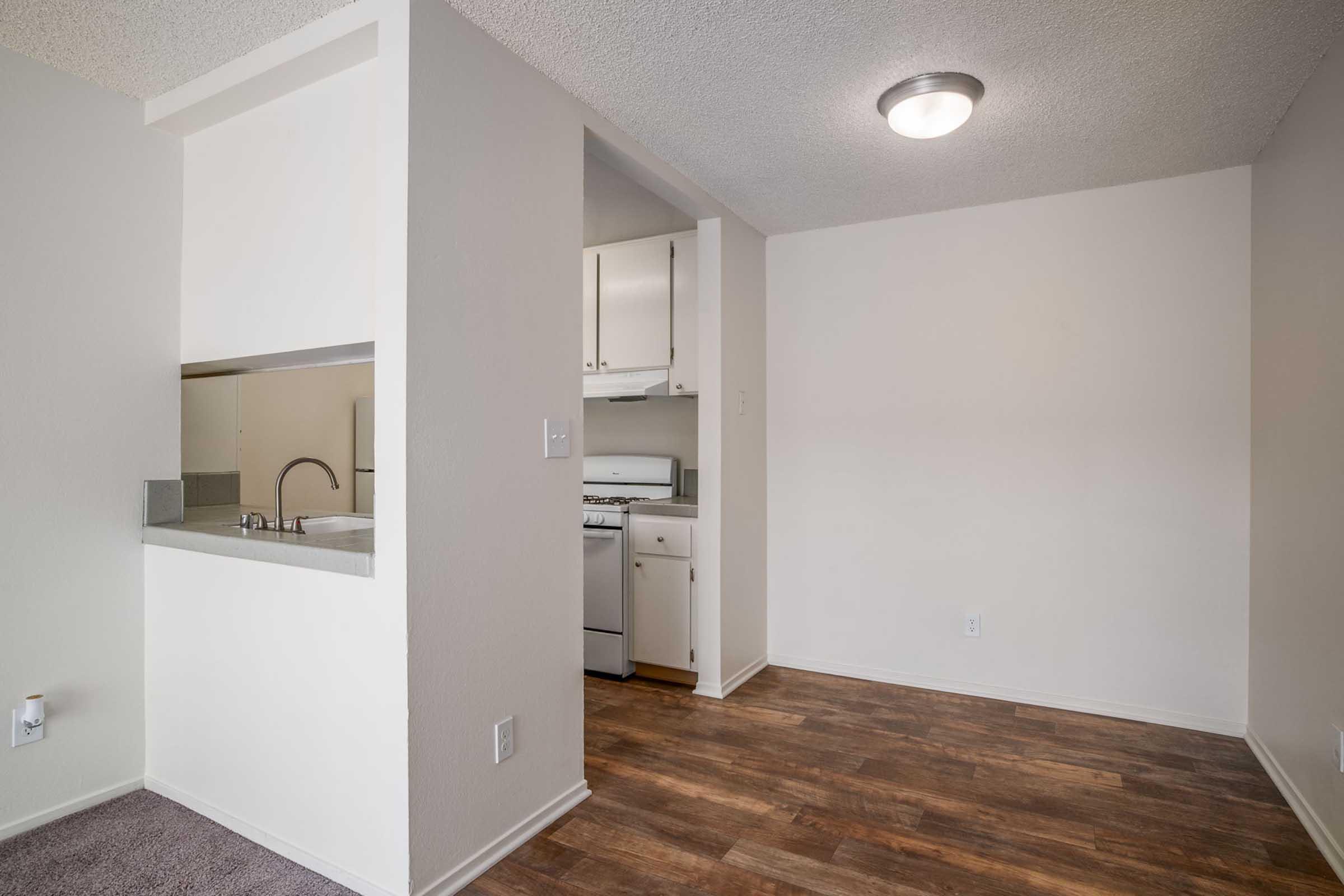 A well-lit room featuring a part of a kitchen with a sink and stove. The wall is partially open to another space, and the flooring is wood-like. The walls are painted a light color, and the ceiling has a simple overhead light fixture. The overall atmosphere is clean and modern, with neutral tones.