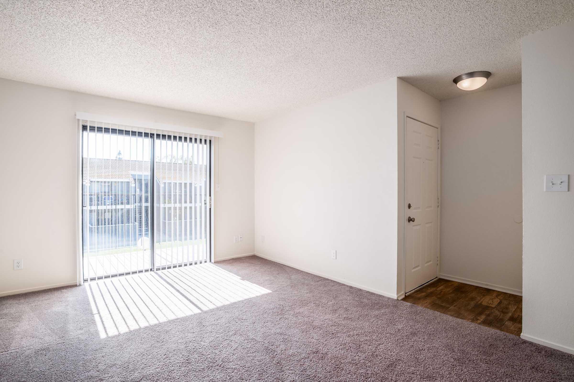 A spacious, brightly lit living room with light beige walls and carpet. Large sliding glass doors lead to an outdoor area, allowing natural light to filter in through vertical blinds. A beige wall separates the living area from a doorway, which features a light fixture above.