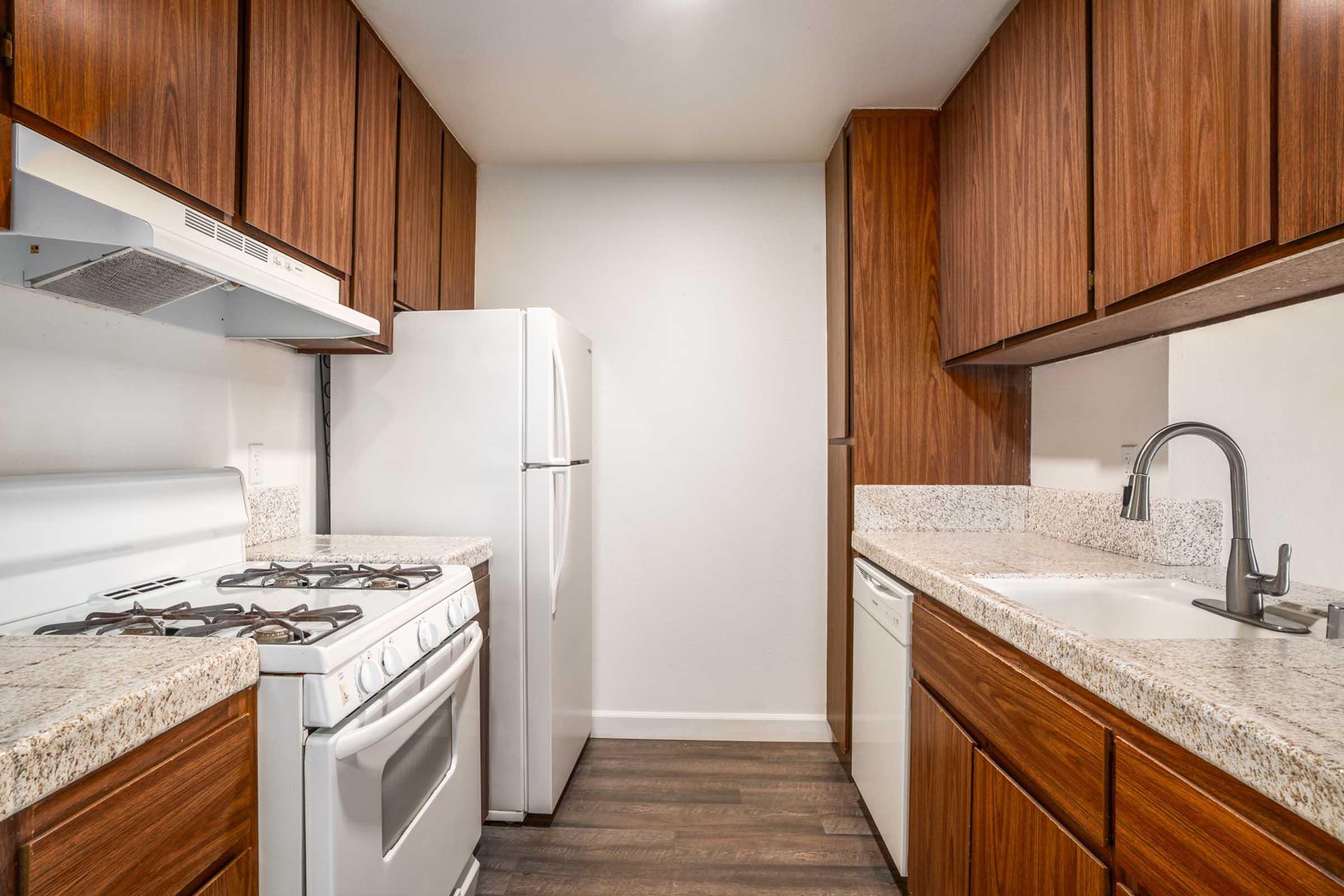 A modern kitchen featuring wooden cabinets, a white gas stove, a refrigerator, and a dishwasher. The countertops are light-colored granite, and the kitchen includes a stainless steel sink with a sleek faucet. The overall design is clean and contemporary.