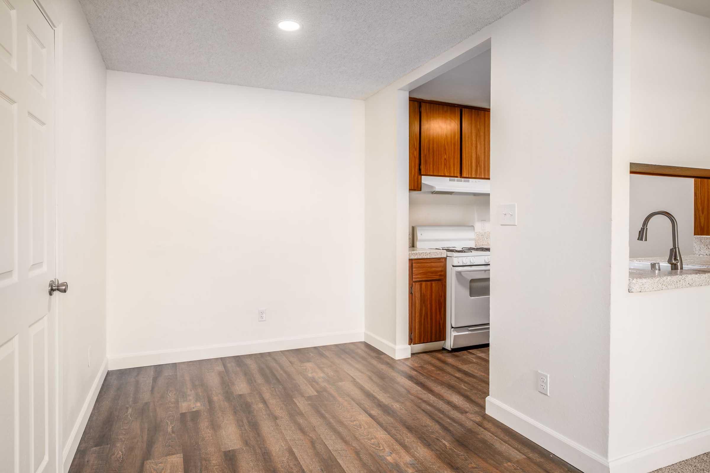 A bright, modern living space featuring a hardwood floor, white walls, and a small kitchen area with wooden cabinets. The kitchen includes a stove and an open layout connecting to a living room. There is a door leading to another room, and ample natural light illuminates the area.