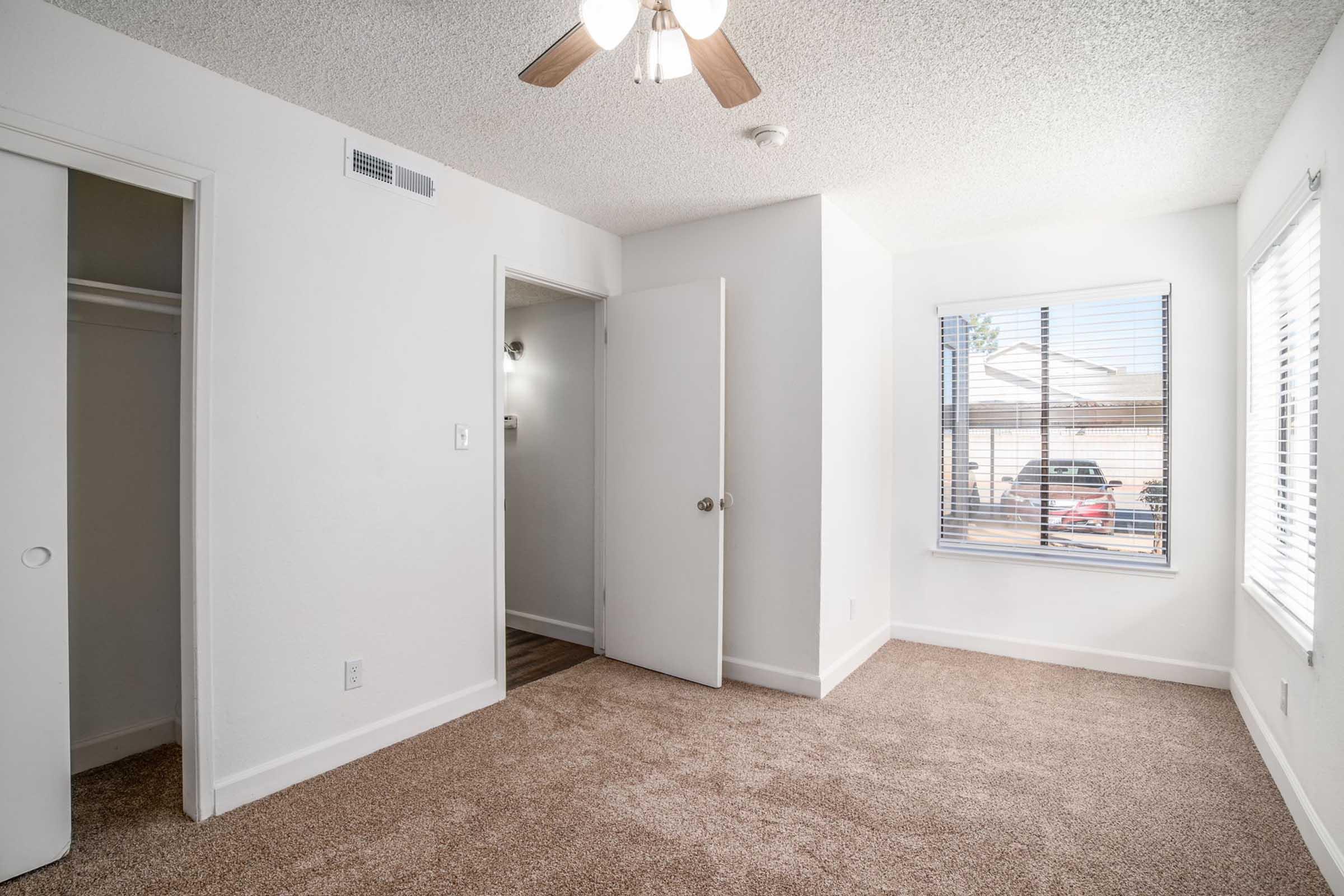 A well-lit bedroom featuring beige carpet, a ceiling fan, and white walls. There is a door leading outside, a closet to the left, and a window with blinds allowing natural light in. The view outside includes vehicles parked in a driveway.