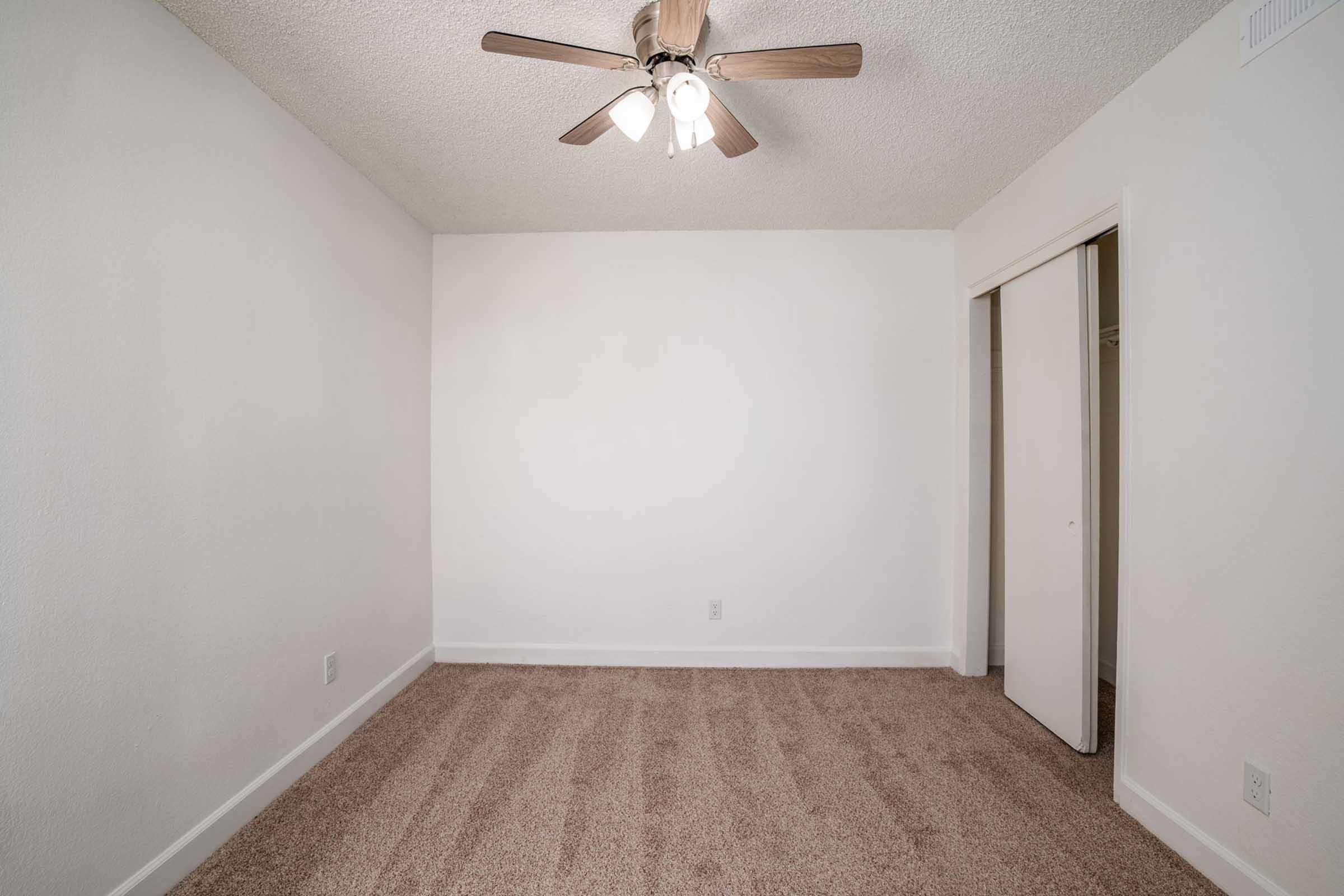 Empty room with light-colored walls and a ceiling fan with wooden blades. The floor is covered in beige carpet. There is a closet space visible on the right side, and the overall ambiance is bright and minimalistic.