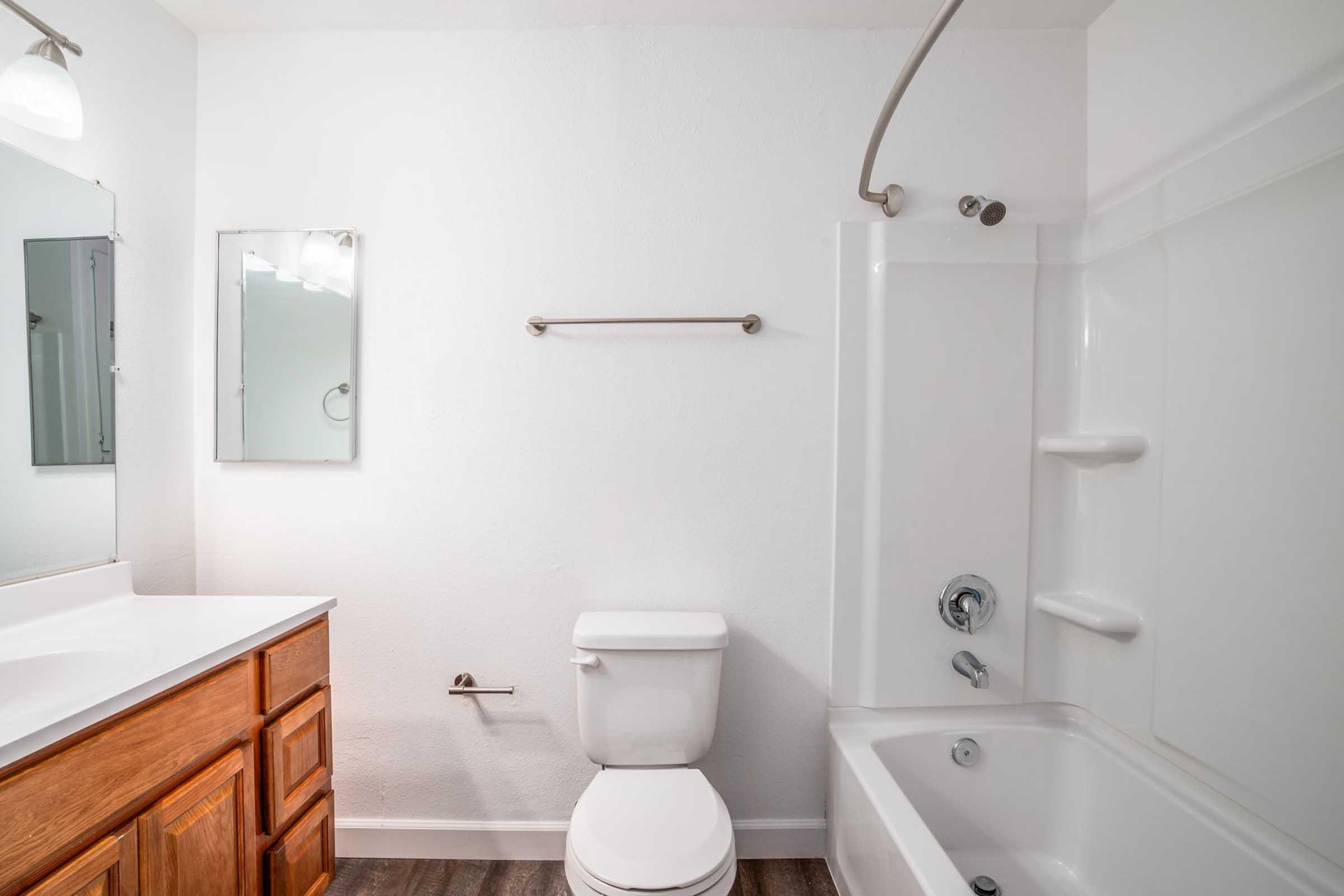A clean and modern bathroom featuring a white bathtub with shower, a toilet, and a vanity with a sink. There are two mirrors on the wall, a towel bar, and neutral-colored walls and flooring, creating a bright and spacious atmosphere.