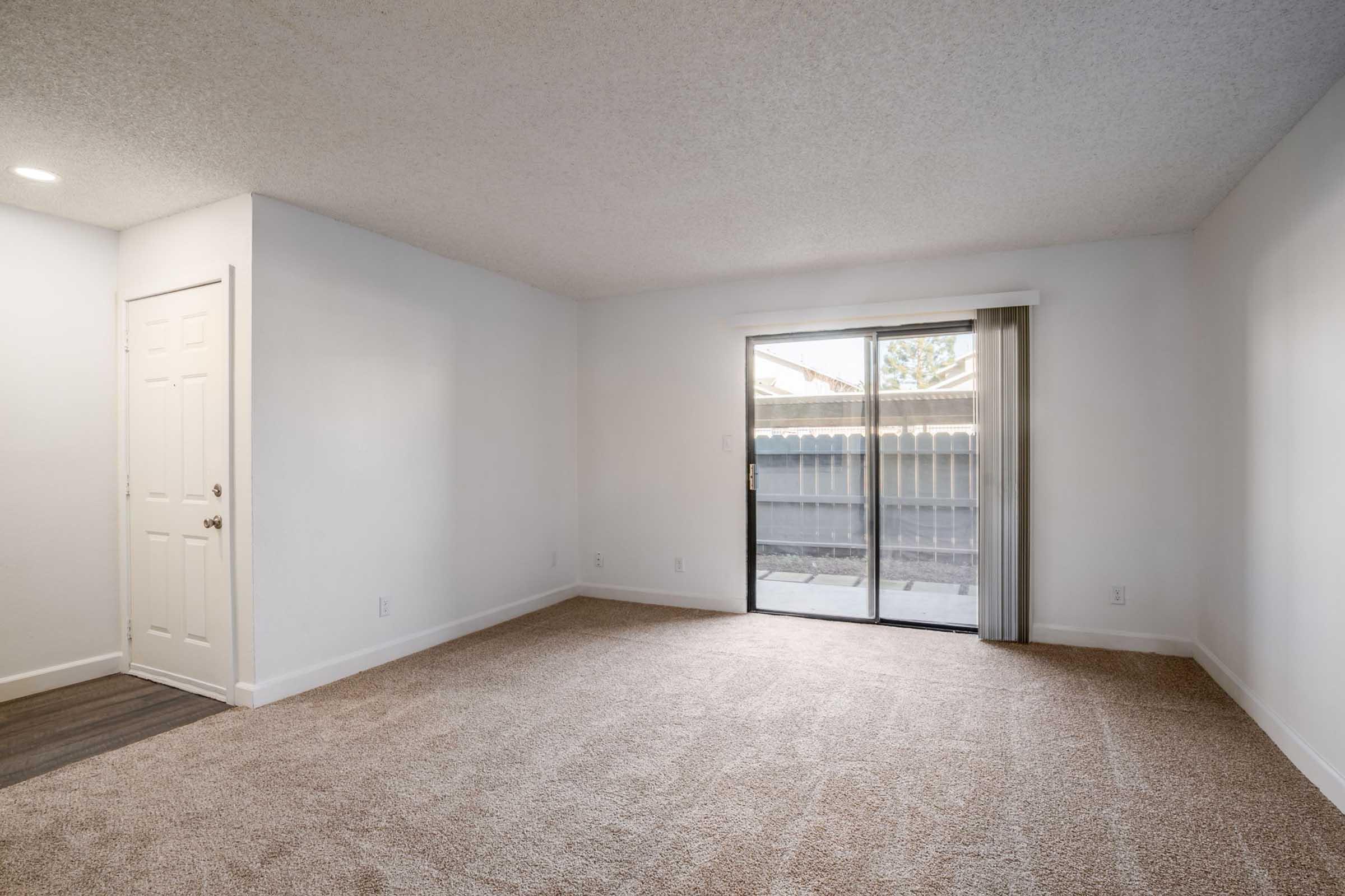 A spacious, empty living room with light beige carpet and white walls. A sliding glass door leads to a small outdoor area, providing natural light. A closed door on the left suggests access to another room. The overall atmosphere is bright and inviting, suitable for various interior designs.