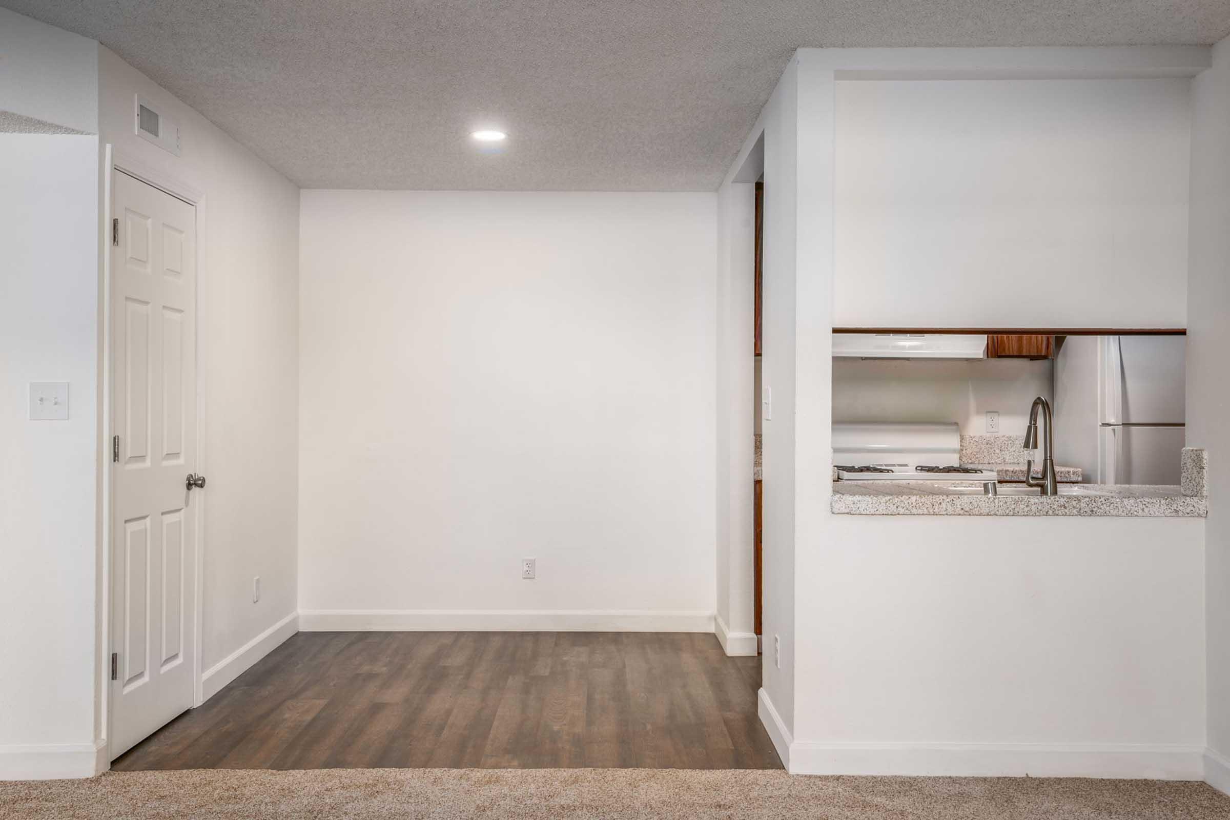A well-lit interior of an apartment featuring a partially open kitchen with a modern countertop, a stainless steel fridge, and a doorway leading to another room. The flooring is a mix of carpet and laminate, with white walls and a simple, clean design.