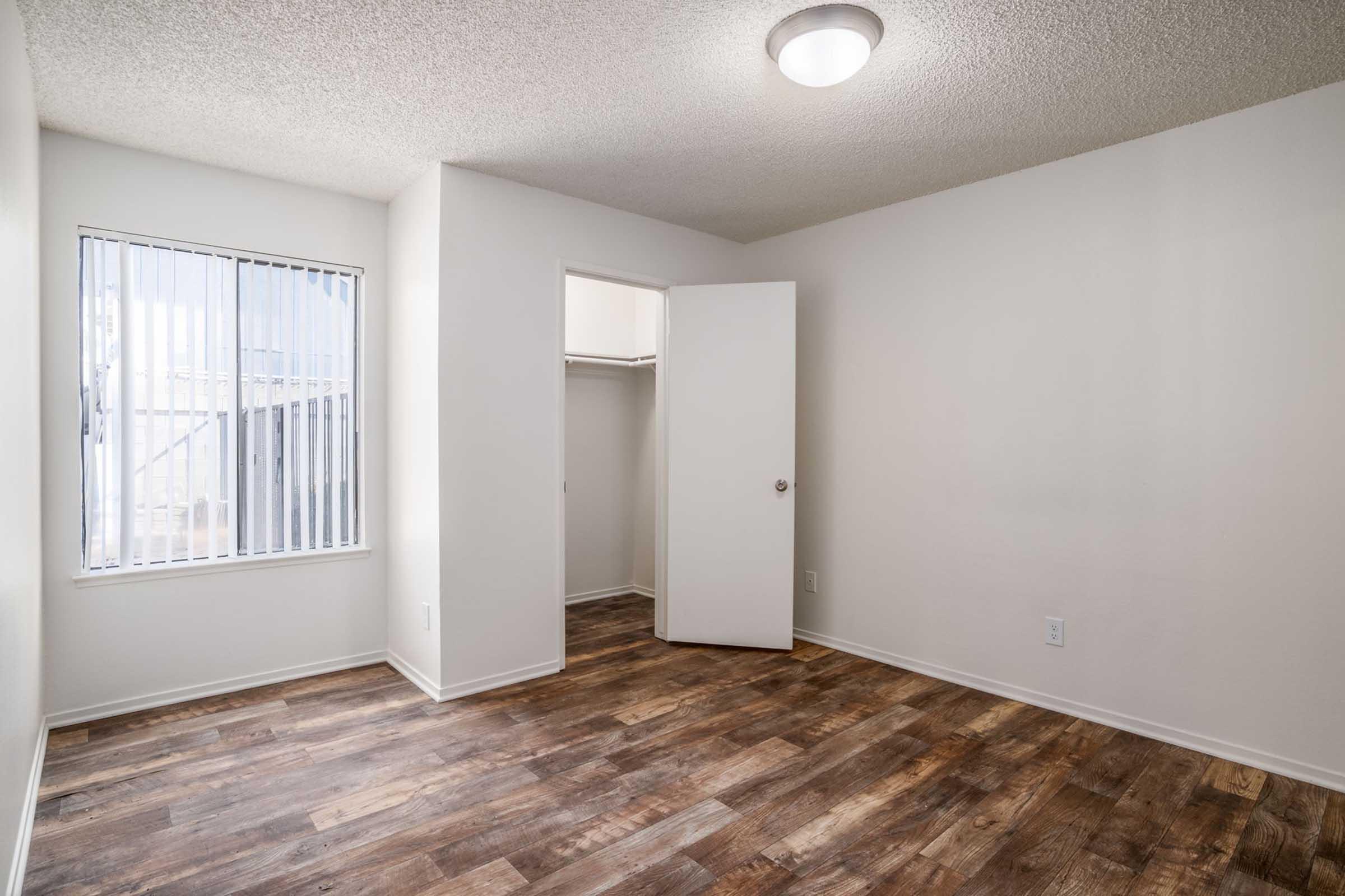 A bright, empty bedroom featuring light-colored walls and a textured ceiling. It has a window with vertical blinds, a closet with an open door, and wooden flooring. The space is well-lit and minimalistic, creating a clean, open atmosphere.