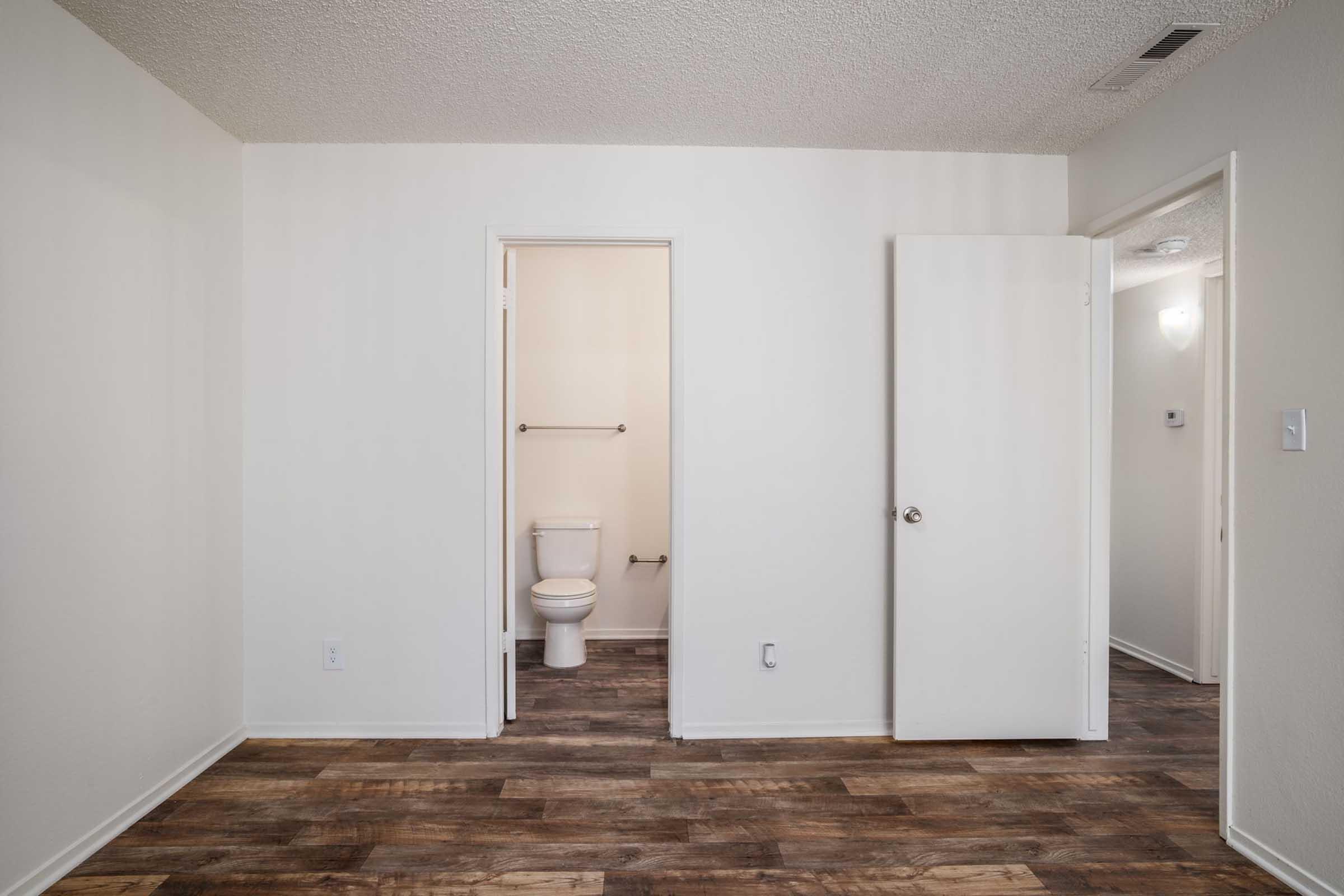 A softly lit empty room featuring light-colored walls and a wood-patterned floor. A doorway leads to a small bathroom with a toilet visible. Another door is partially ajar, suggesting access to another space. The overall atmosphere is clean and simple, ideal for a living or bedroom area.