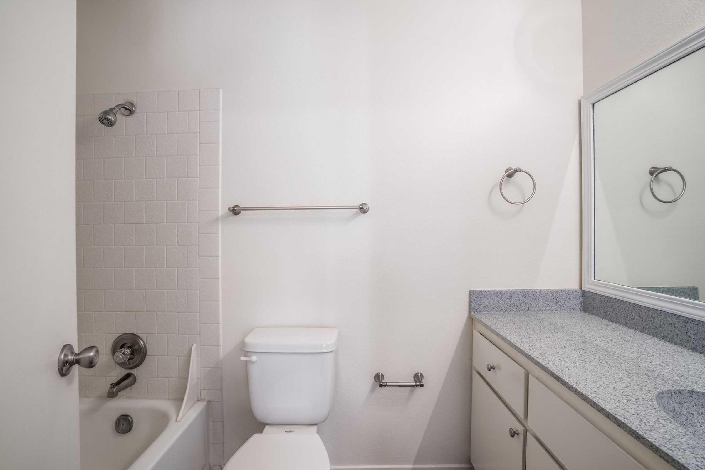 A clean and modern bathroom featuring a bathtub with a showerhead, a white toilet, a granite countertop with cabinets, and a wall-mounted mirror. The walls are painted in light colors, and there are towel bars and fixtures visible, creating a simple and functional space.