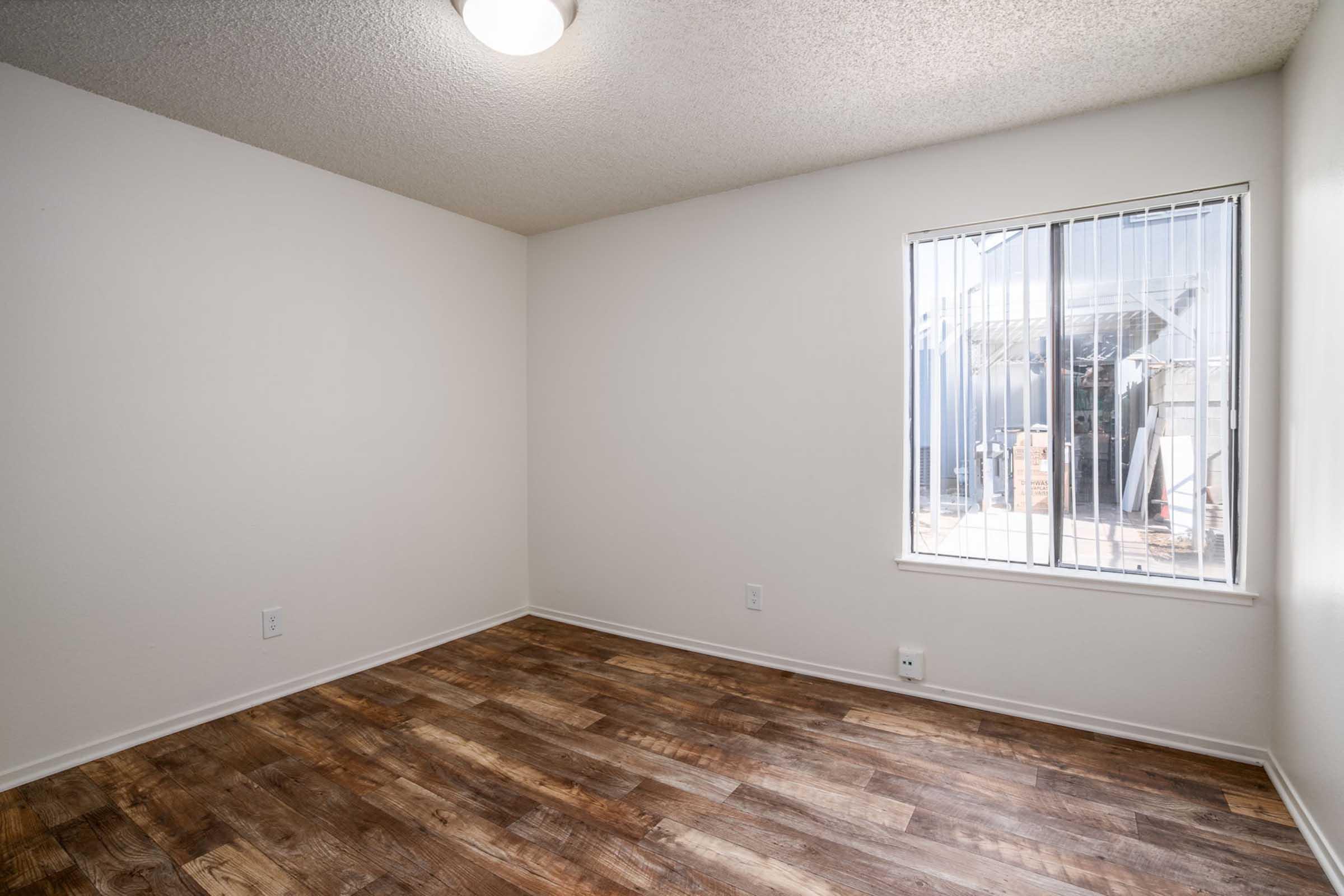 Empty room featuring light-colored walls, a window with vertical blinds, and wooden flooring. The room has a ceiling light fixture and is uncluttered, providing a spacious feel. Outside the window, a glimpse of another building can be seen.