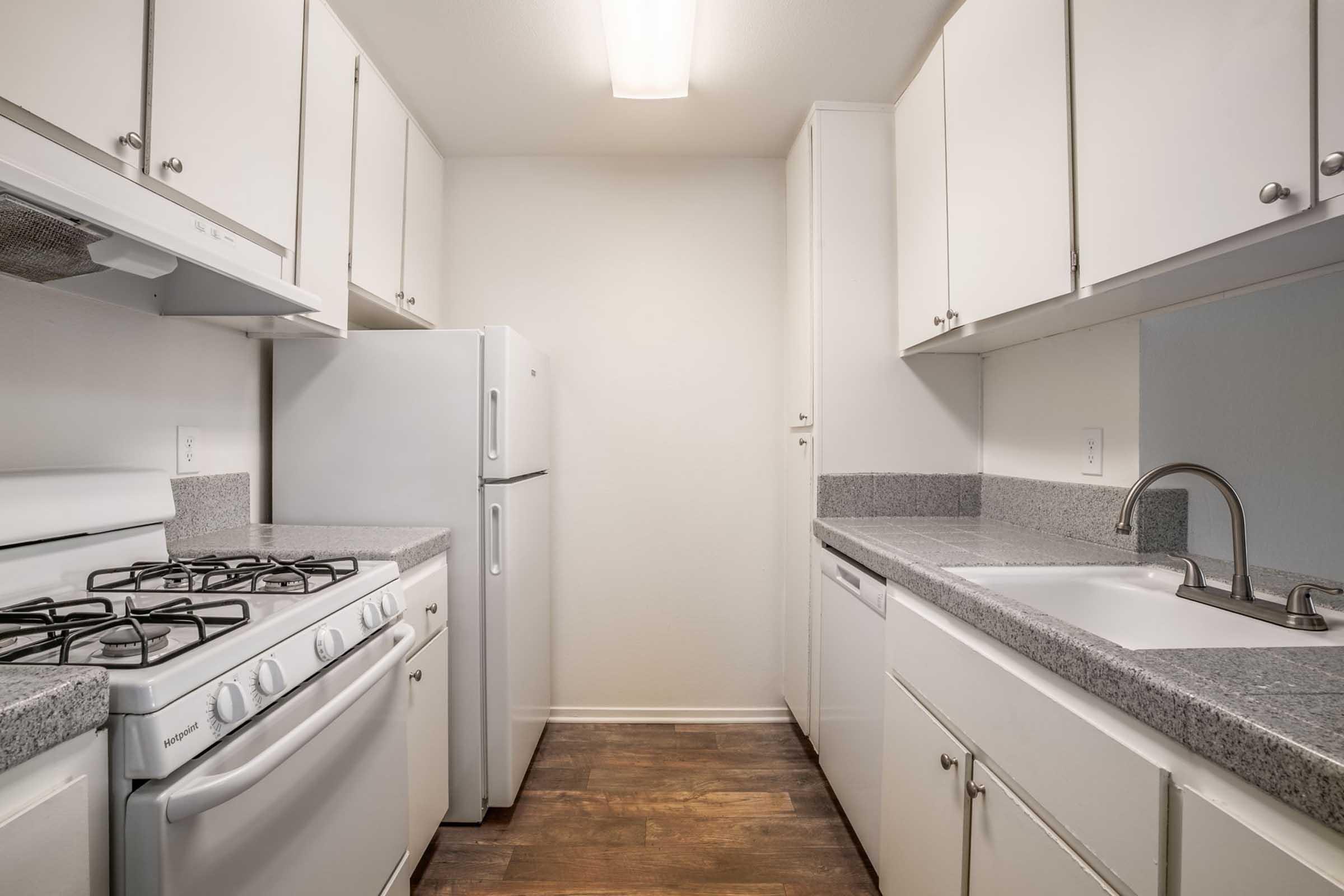 A modern kitchen featuring white cabinetry, a gas stove, a refrigerator, and a sink. The countertops are gray, and the floors appear to be dark wood. The layout is compact, with ample storage and natural light from overhead lighting.