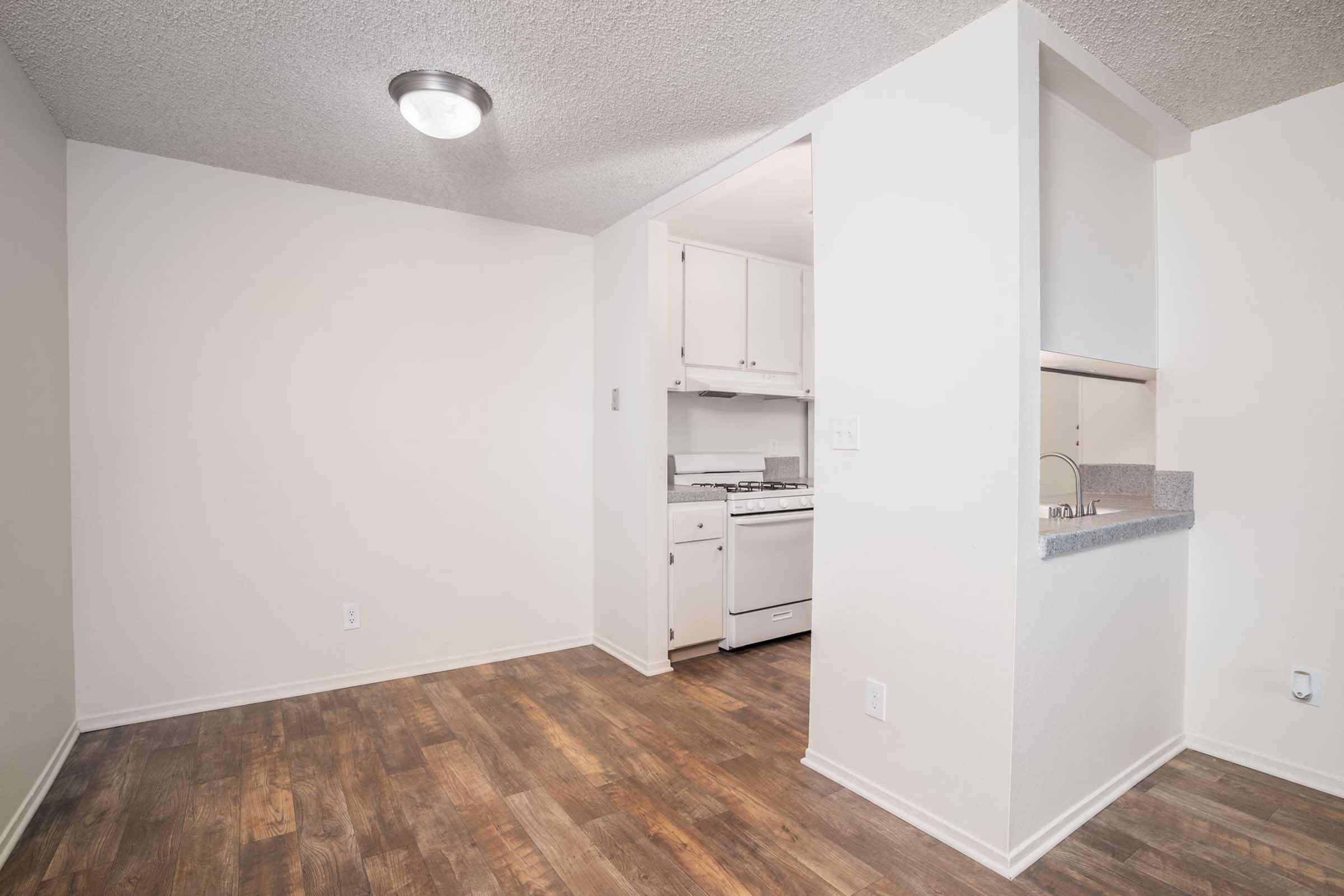 A bright, empty room with beige walls and a ceiling light. To the left, there is a partial view of a compact kitchen featuring white cabinets, a stove, and a sink. The flooring is brown laminate, and a small cut-out connects the kitchen to the main room, creating an open feel.