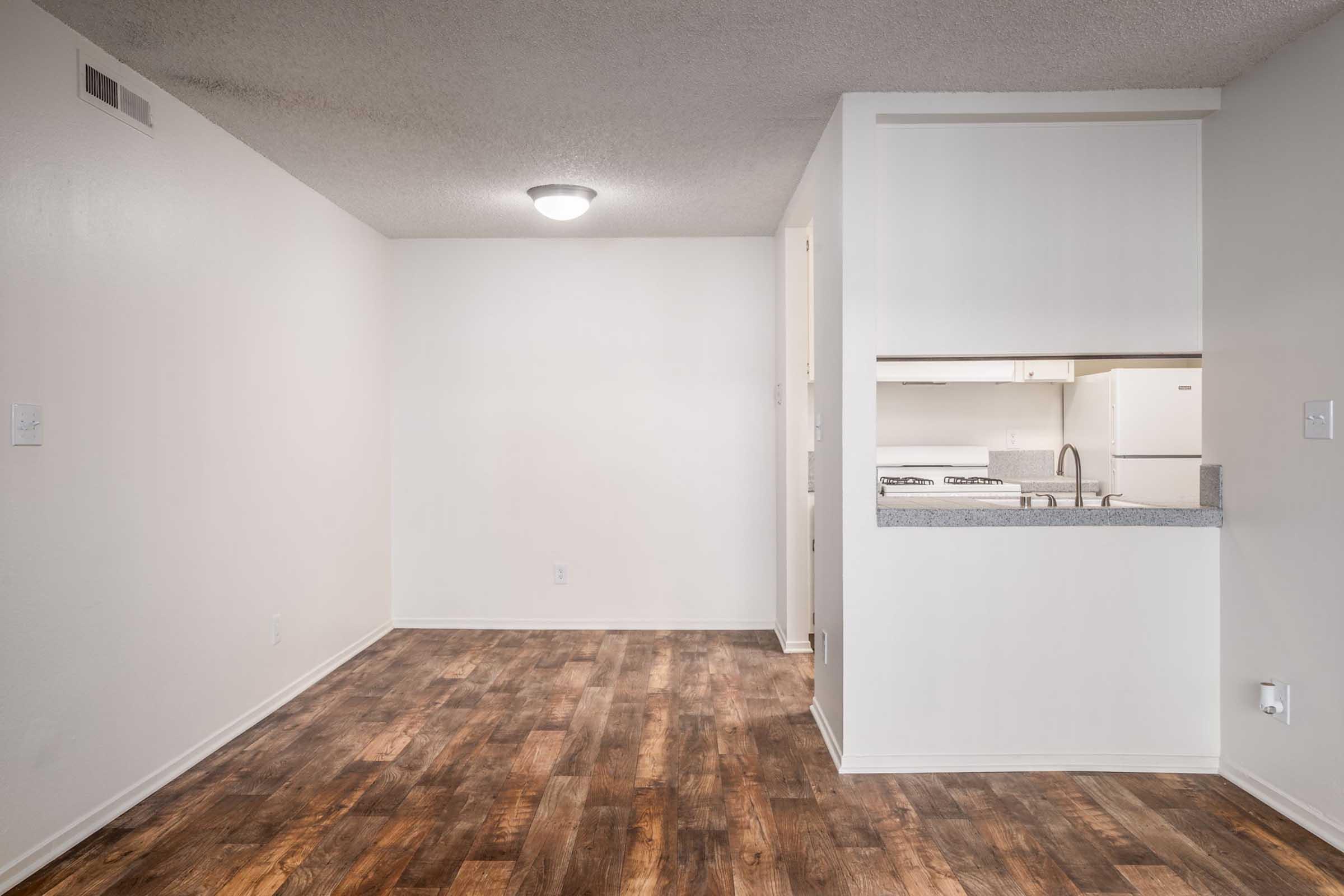 A small, vacant apartment interior featuring light-colored walls, a round ceiling light, and a kitchen area with white appliances visible through an open doorway. The flooring is a wood-like laminate. The space is unoccupied, creating a clean and minimalist look.