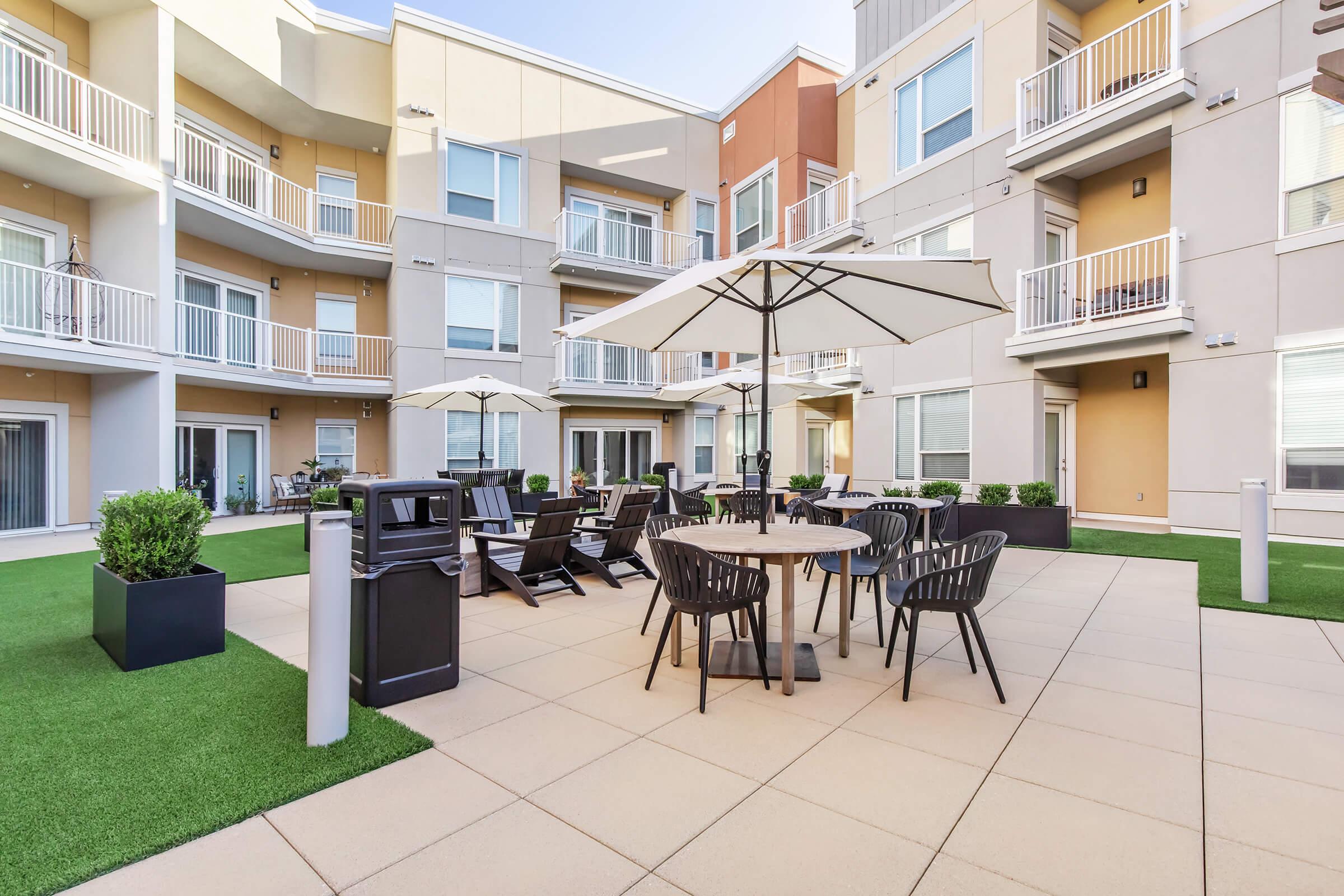 A modern outdoor courtyard featuring a paved area with a round table surrounded by black chairs, umbrellas, and green landscaping. Surrounding the courtyard are several apartment buildings with balconies, creating a welcoming and communal atmosphere.