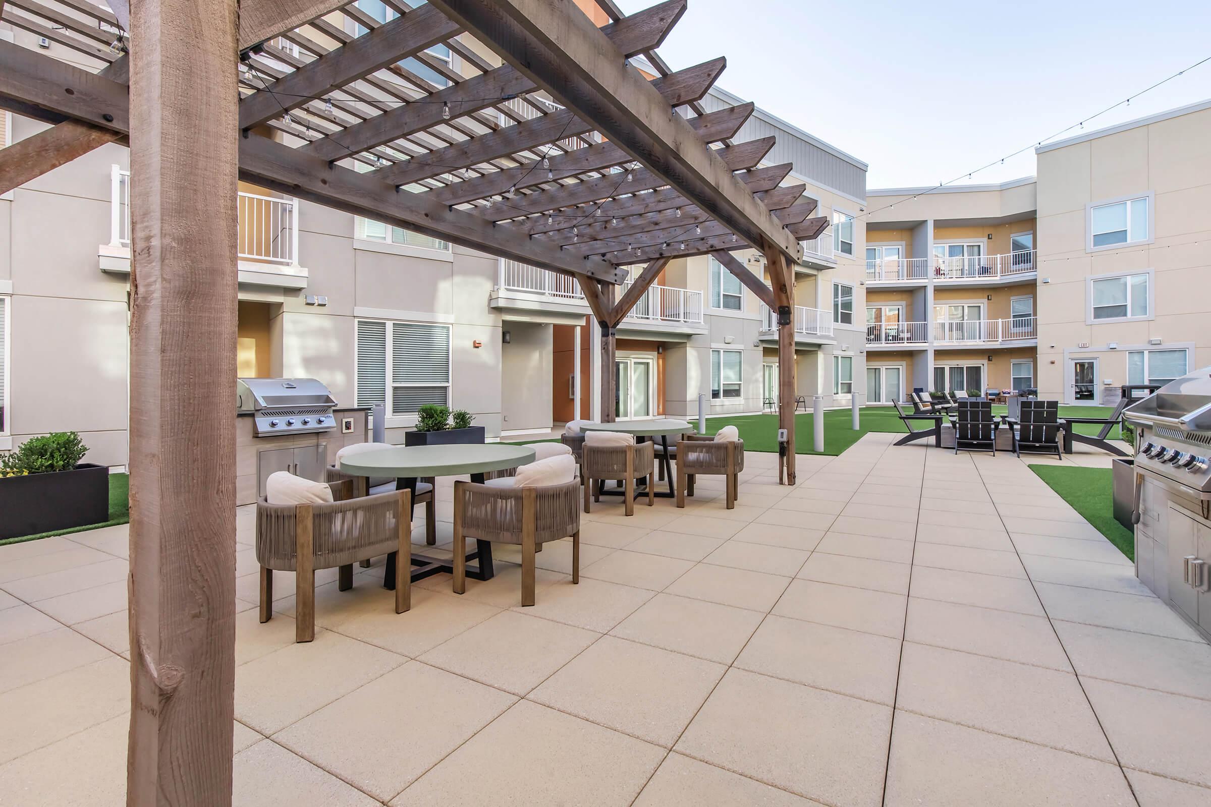 Outdoor communal area featuring a pergola, dining table, and chairs on a tiled patio. In the background, several apartment units are visible, along with a barbecue grill. The space is designed for relaxation and social gatherings, surrounded by green grass.