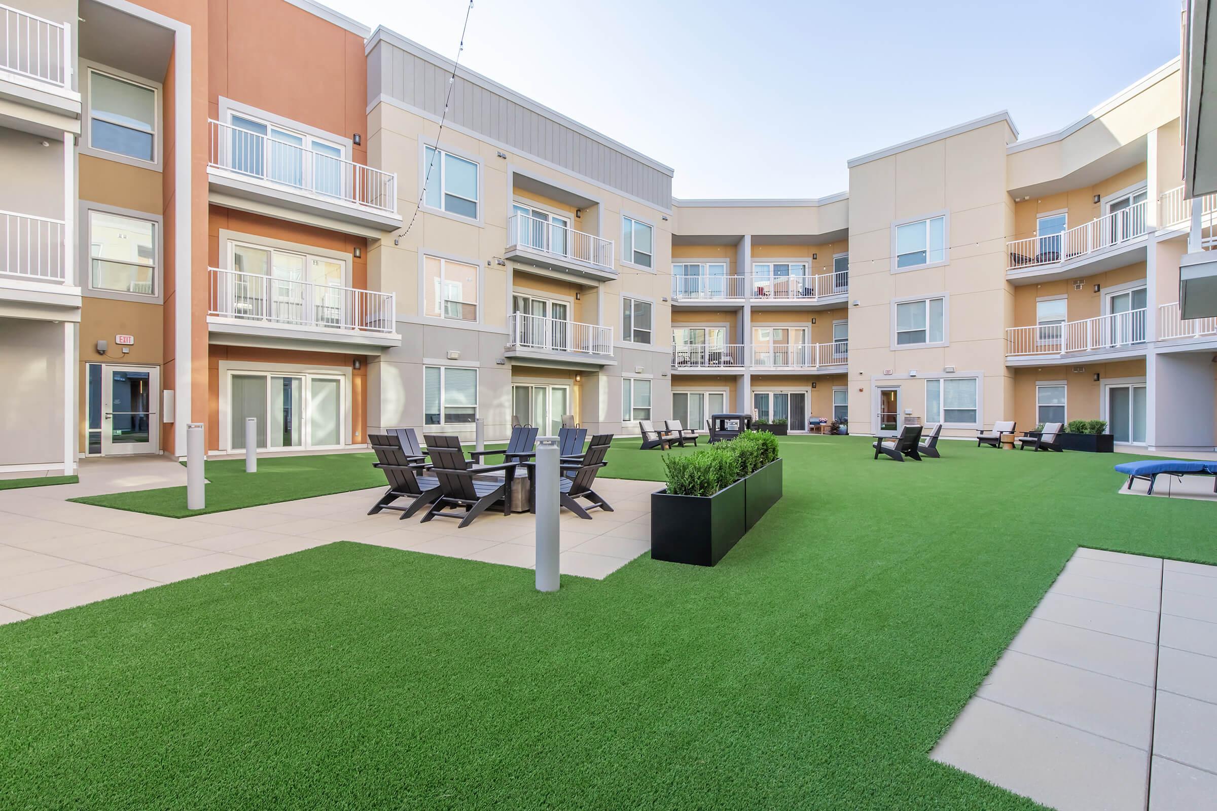 A modern apartment courtyard featuring green artificial turf, sleek lounge chairs, and contemporary buildings with balconies. The space is well-lit and inviting, ideal for outdoor gatherings and relaxation.