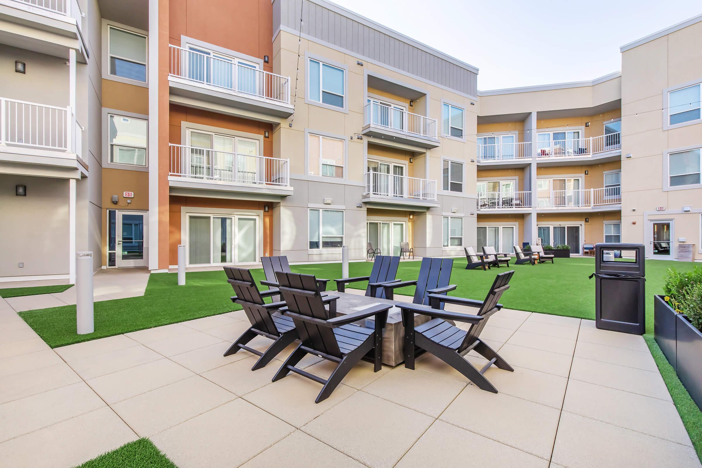 A well-maintained outdoor area featuring modern apartment buildings with balconies. In the foreground, there is a seating area with black wooden chairs and a table, surrounded by manicured grass. The surroundings include additional lounge chairs and a trash bin, creating a welcoming communal space.