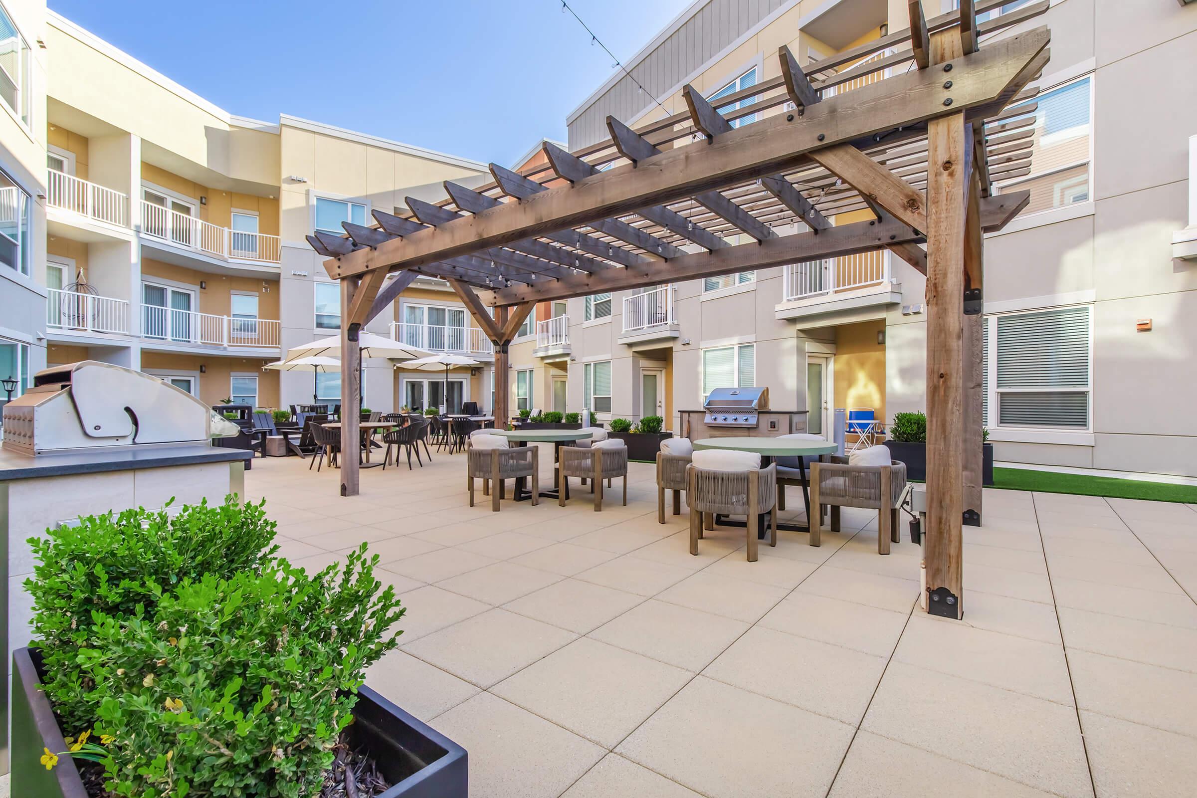 Outdoor patio area featuring a wooden pergola, tables, and chairs. There is a barbecue grill under the pergola, surrounded by well-maintained landscaping. The setting is part of a residential complex with modern buildings in the background and a clear blue sky overhead.
