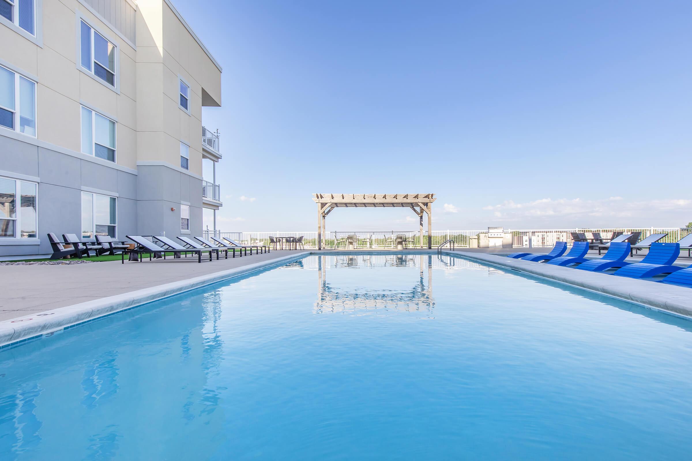 A serene outdoor swimming pool with crystal-clear water, surrounded by lounge chairs on a sunny day. In the background, a wooden pergola adds a decorative touch, while the sky is bright blue with few clouds, creating a relaxing atmosphere.