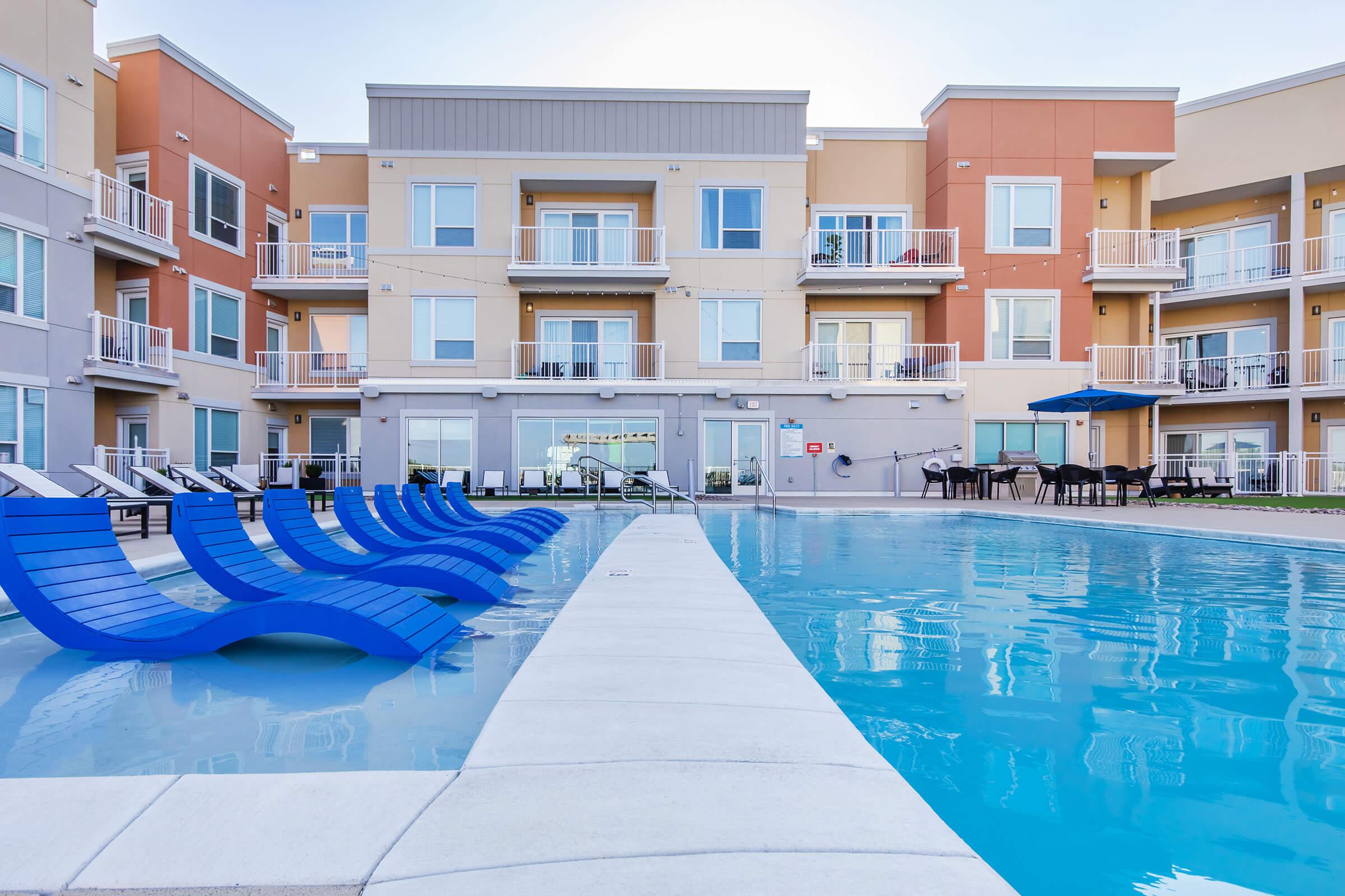 A modern apartment complex featuring a swimming pool with blue lounge chairs lined along the edge. The building has multiple balconies and large windows, with a patio area visible in the background. Clear skies enhance the bright and inviting atmosphere of the outdoor space.