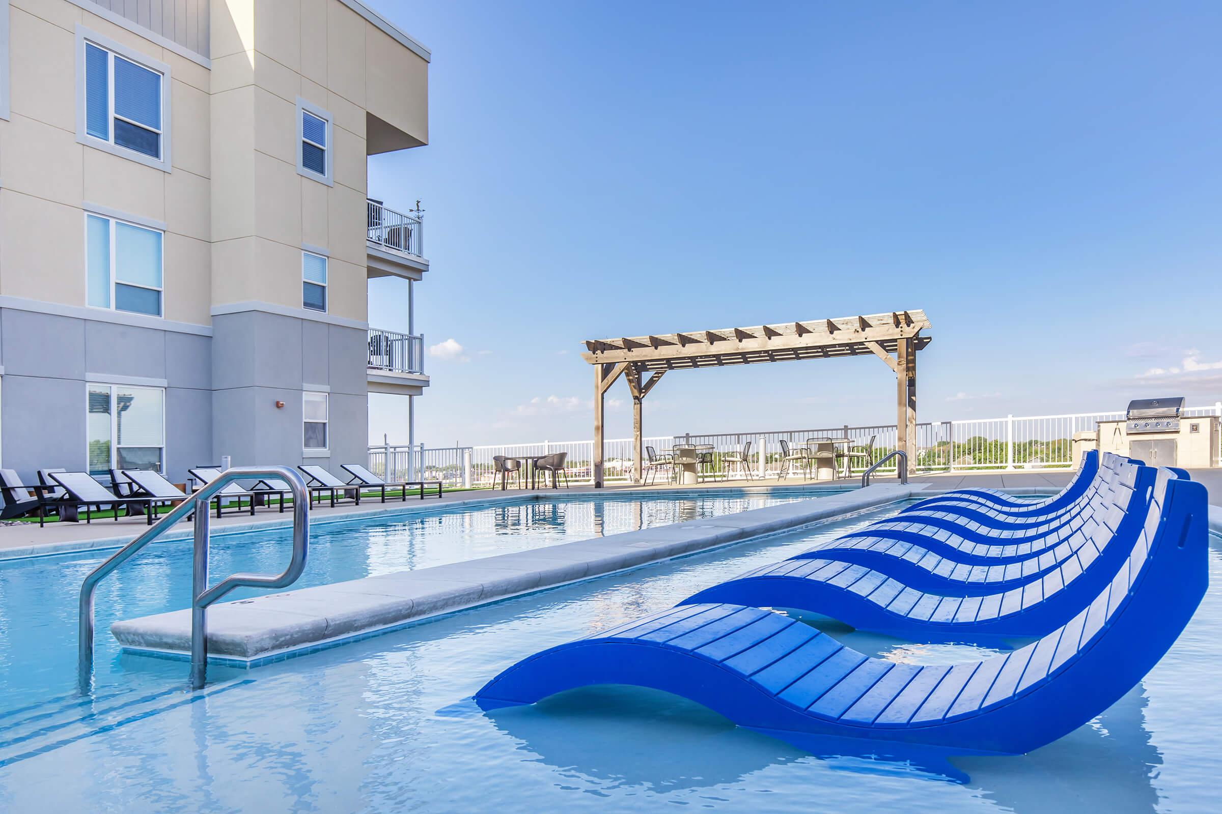 A modern outdoor pool area featuring a long, narrow pool with blue lounge chairs partially submerged in the water. There are white sun loungers along the poolside and a wooden pergola in the background. The sky is clear and blue, creating a serene atmosphere.