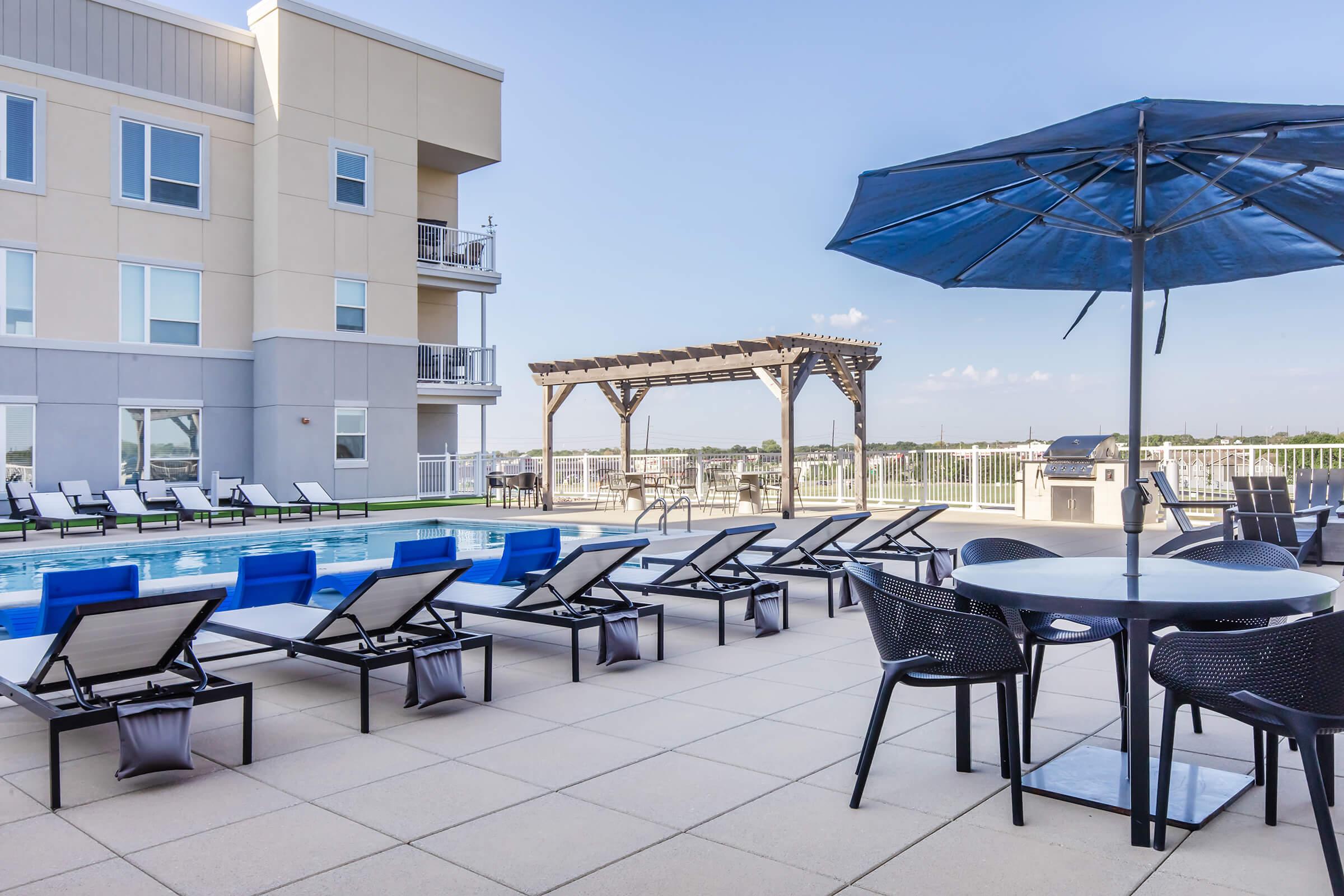 A rooftop pool area featuring lounge chairs and umbrellas, with a nearby pergola. In the background, a multi-story building is visible under a clear blue sky. The pool reflects the surroundings, creating a relaxing outdoor space for leisure.