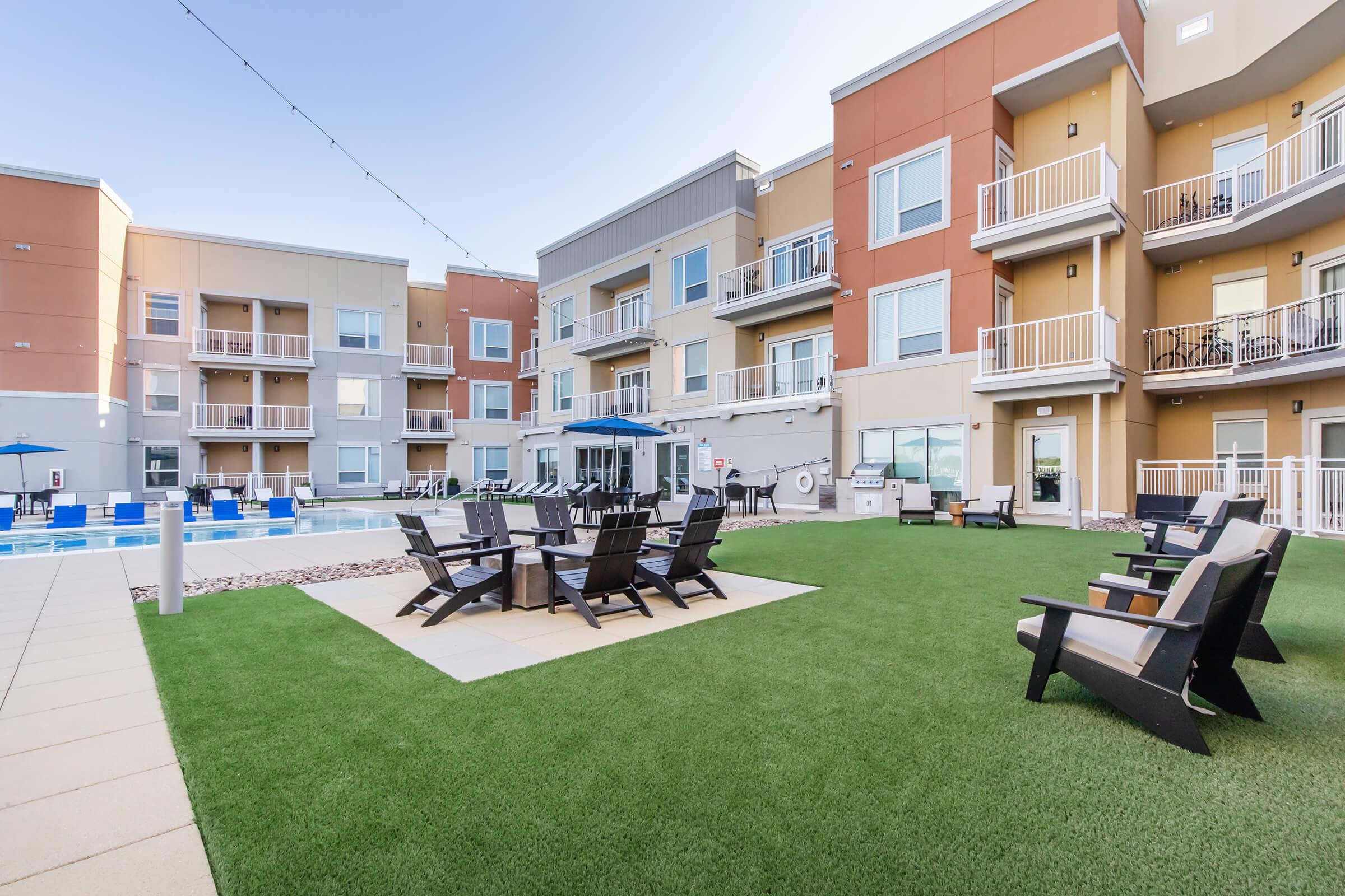 A modern apartment complex featuring a well-maintained pool area surrounded by lounge chairs. In the foreground, there are several black outdoor chairs arranged around a small table on a green lawn, with buildings in the background. The setting is bright and inviting, ideal for relaxation and socializing.