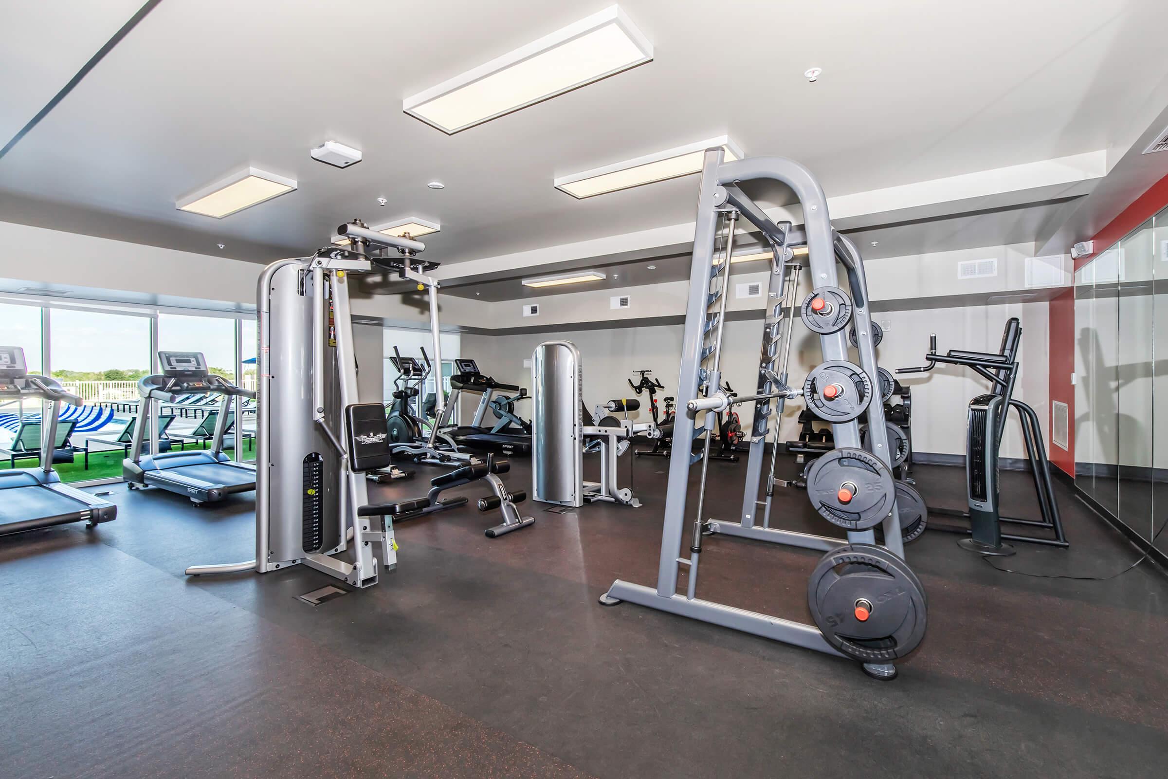 A modern gym interior featuring various fitness equipment, including weight machines, treadmills, and stationary bikes. The space is well-lit with large windows providing natural light, and the floors are covered with dark rubber flooring for safety and comfort.