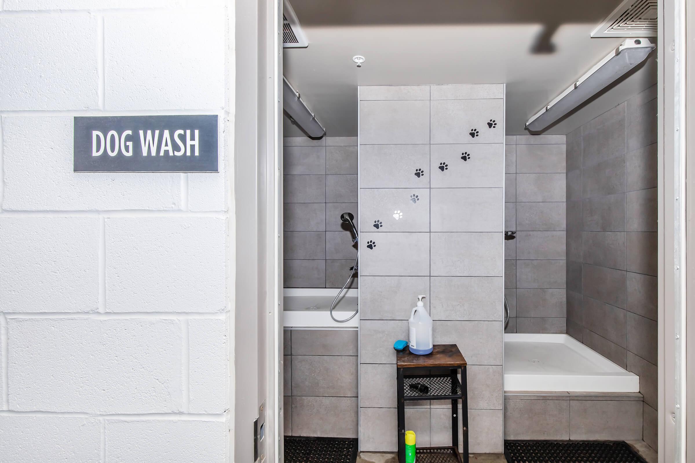 A self-service dog wash station with two bathtubs, featuring a tiled wall decorated with paw prints. A small table holds cleaning supplies like shampoo and a spray bottle. A sign above the entrance reads "DOG WASH." The space is clean and designed for washing dogs comfortably.