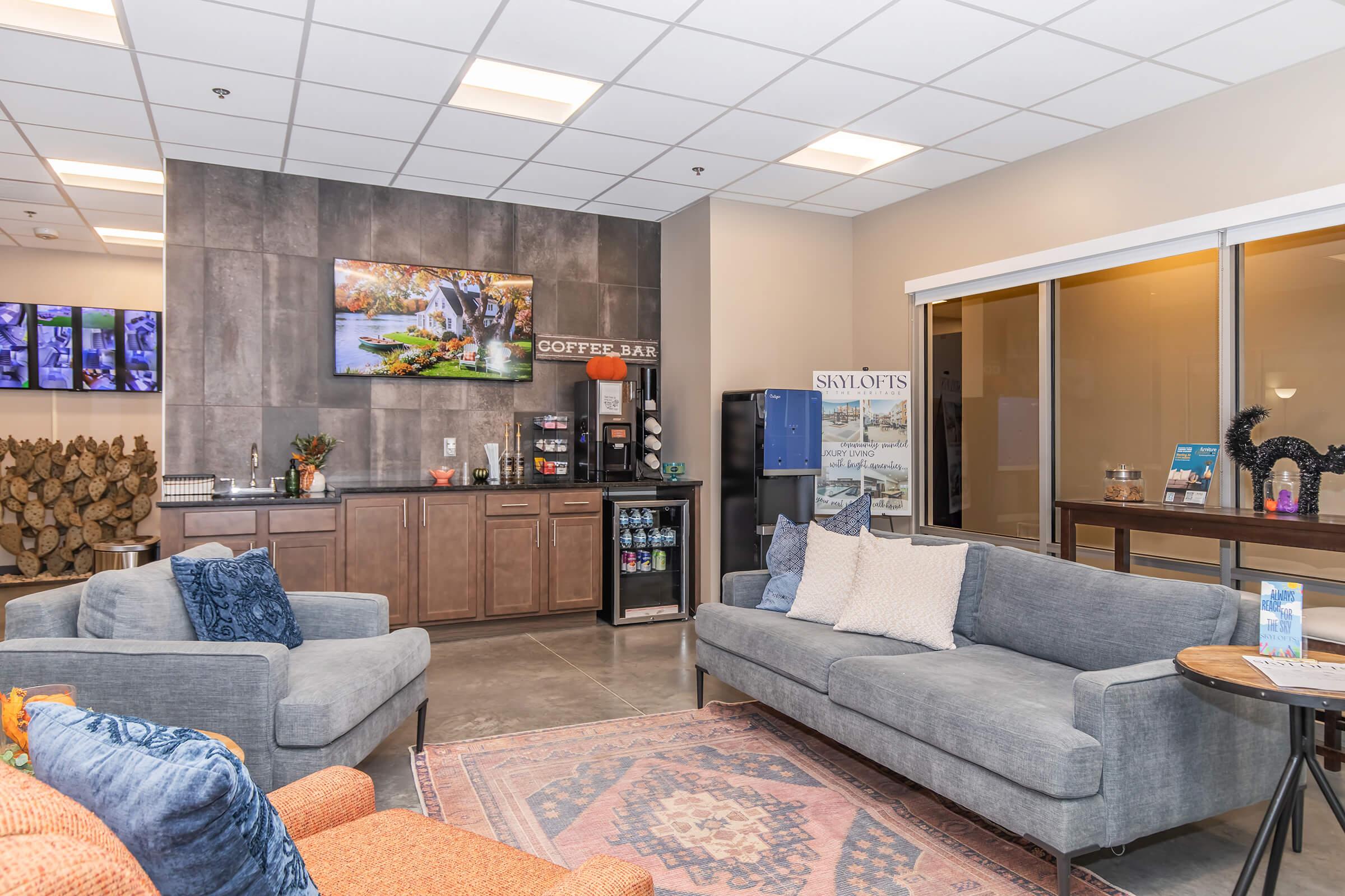 A cozy lounge area featuring two sofas, a coffee bar with various beverages, and a decorative wall-mounted painting. There is a water dispenser and a display sign, along with a patterned area rug on the concrete floor. Natural light enters through large windows, creating a welcoming atmosphere.