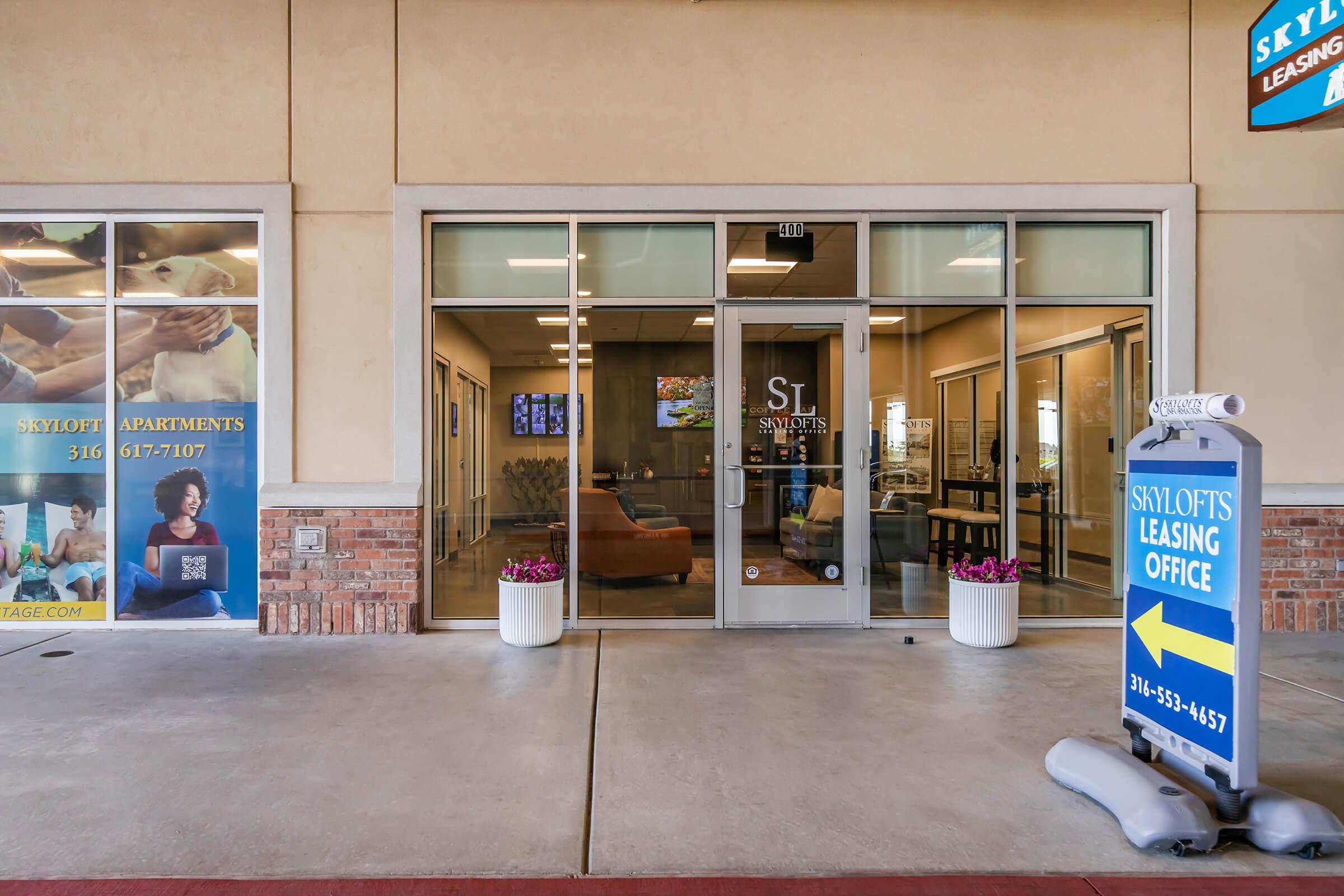 Leasing office entrance with glass doors and a sign, showcasing the Skyl lofts. The area is well-lit, featuring seating visible through the window. Flower pots are placed at the entrance, and promotional banners for apartments are displayed on the side.