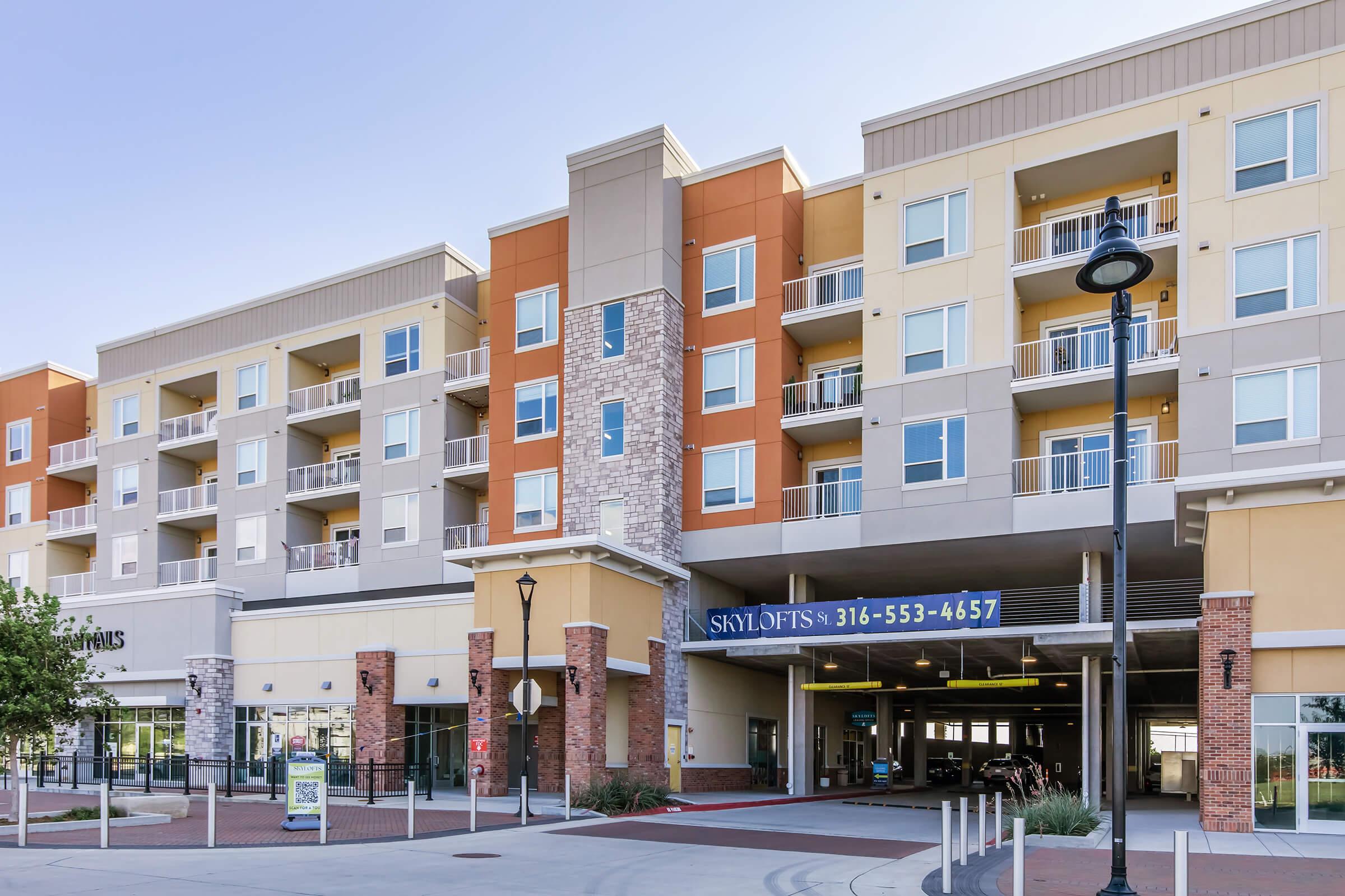 Modern apartment building with a combination of orange, beige, and gray exterior. The ground floor features a parking entrance with a sign for "SkyLofts" displaying a phone number. There are balconies visible on upper floors, with landscaping and street lamps in the foreground.