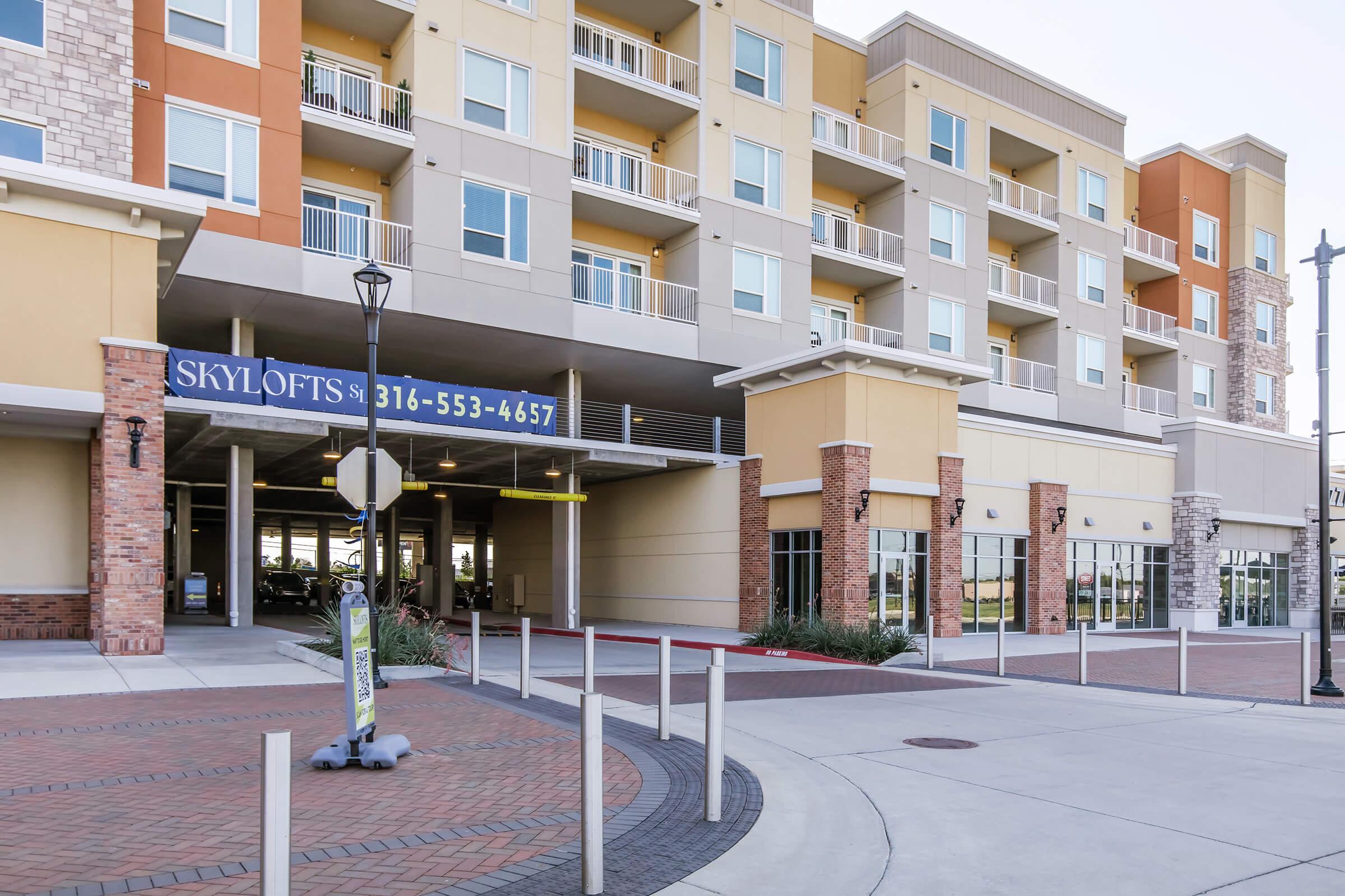 Exterior view of a modern apartment building with multiple stories. The entrance features a sign for "SkyLofts" and a phone number. There are landscaped areas and a circular driveway in front, along with designated parking spaces. The building has a mix of brick and colorful siding.
