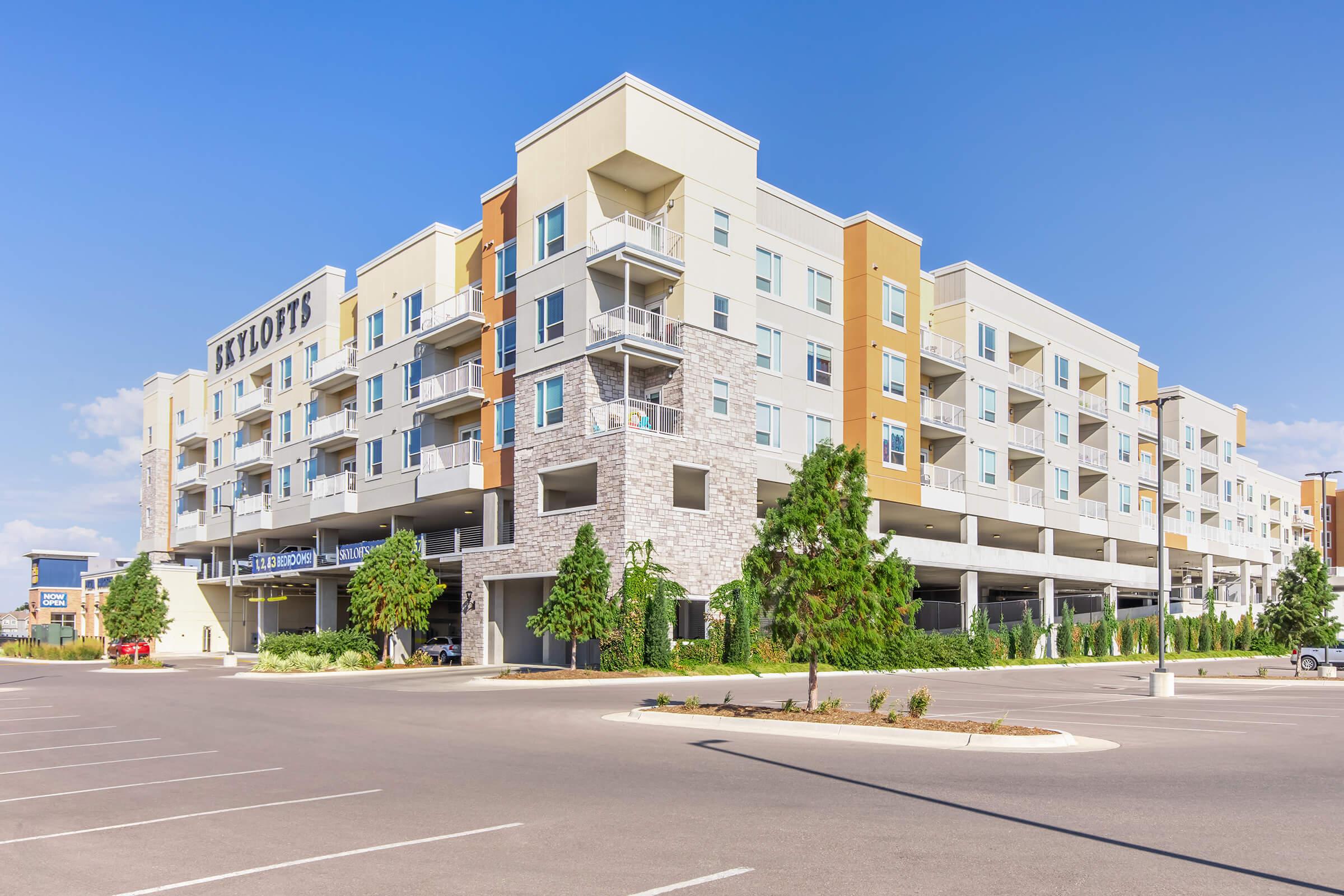 Modern multi-story apartment building with a mix of beige and orange accents, featuring balconies and large windows. The structure includes a covered parking area at the ground level. In the foreground, a spacious parking lot is visible, and the sky is clear and blue.