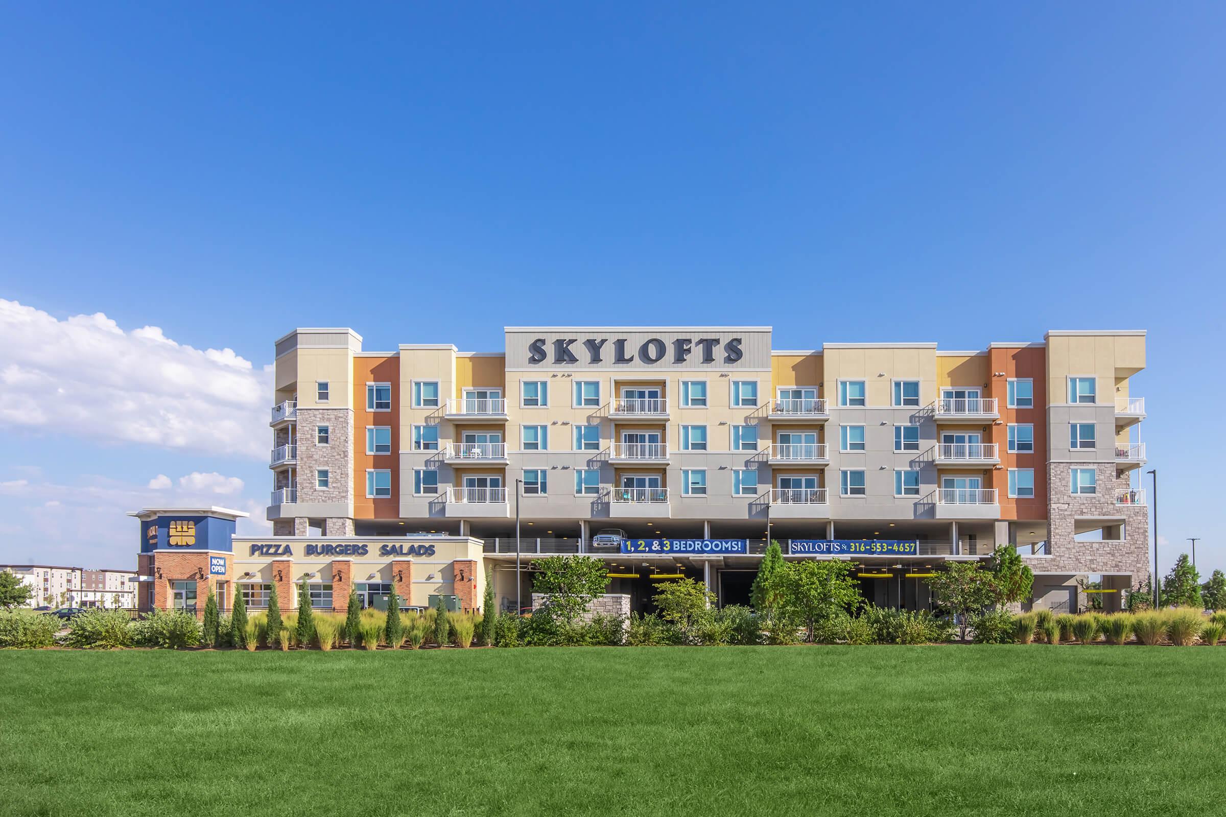 A modern multi-story building named "SKYLOFTS" with a variety of colorful balconies. The ground floor features signage for a restaurant serving pizza, burgers, and salads. Green grassy area in the foreground, with a clear blue sky and scattered clouds in the background.