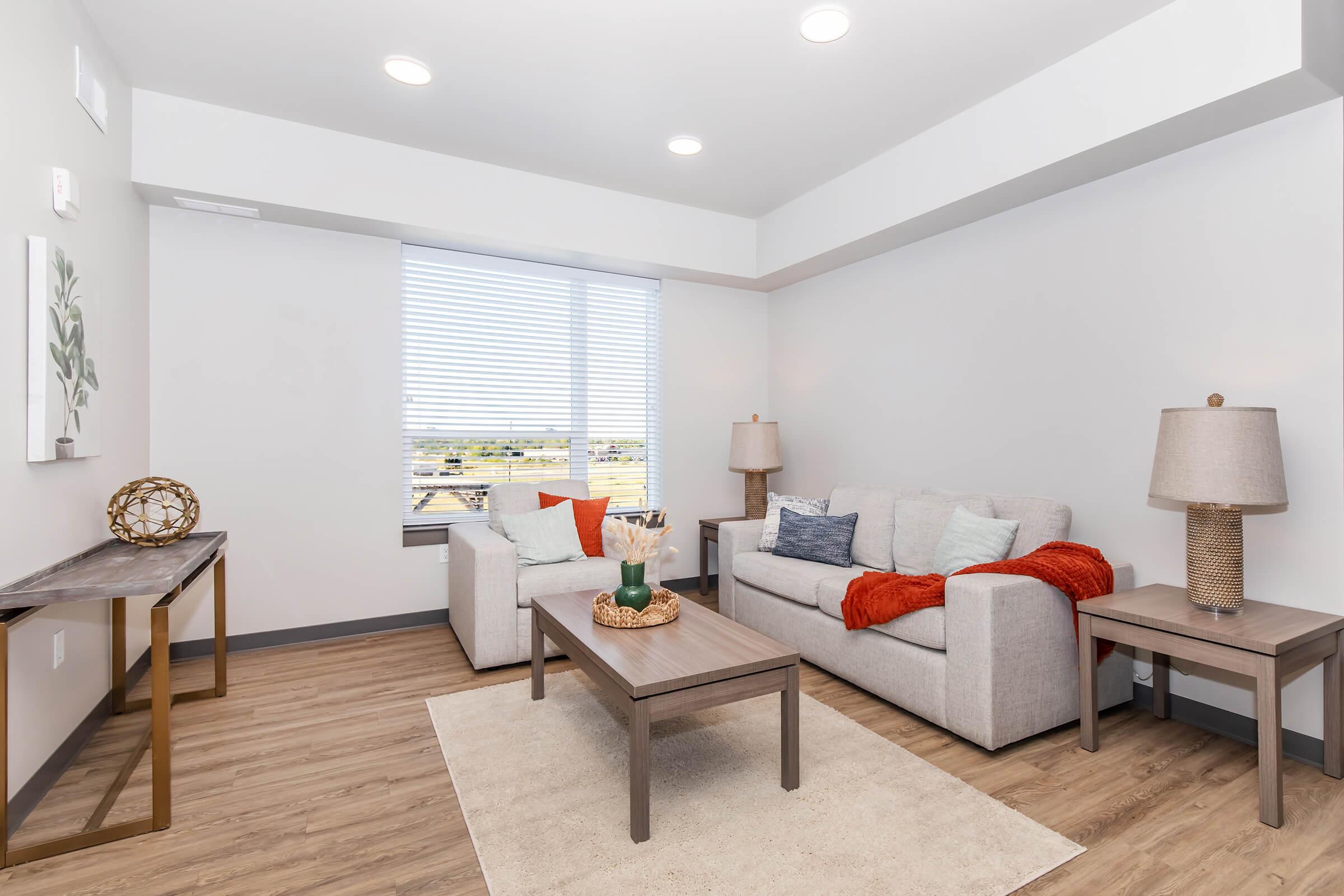 A modern living room featuring a light-colored sofa with decorative cushions, a white armchair, and a wooden coffee table. There are lamps on either side, a textured area rug on the floor, and a decorative bowl on the table. Large windows provide natural light and a view to the outside.